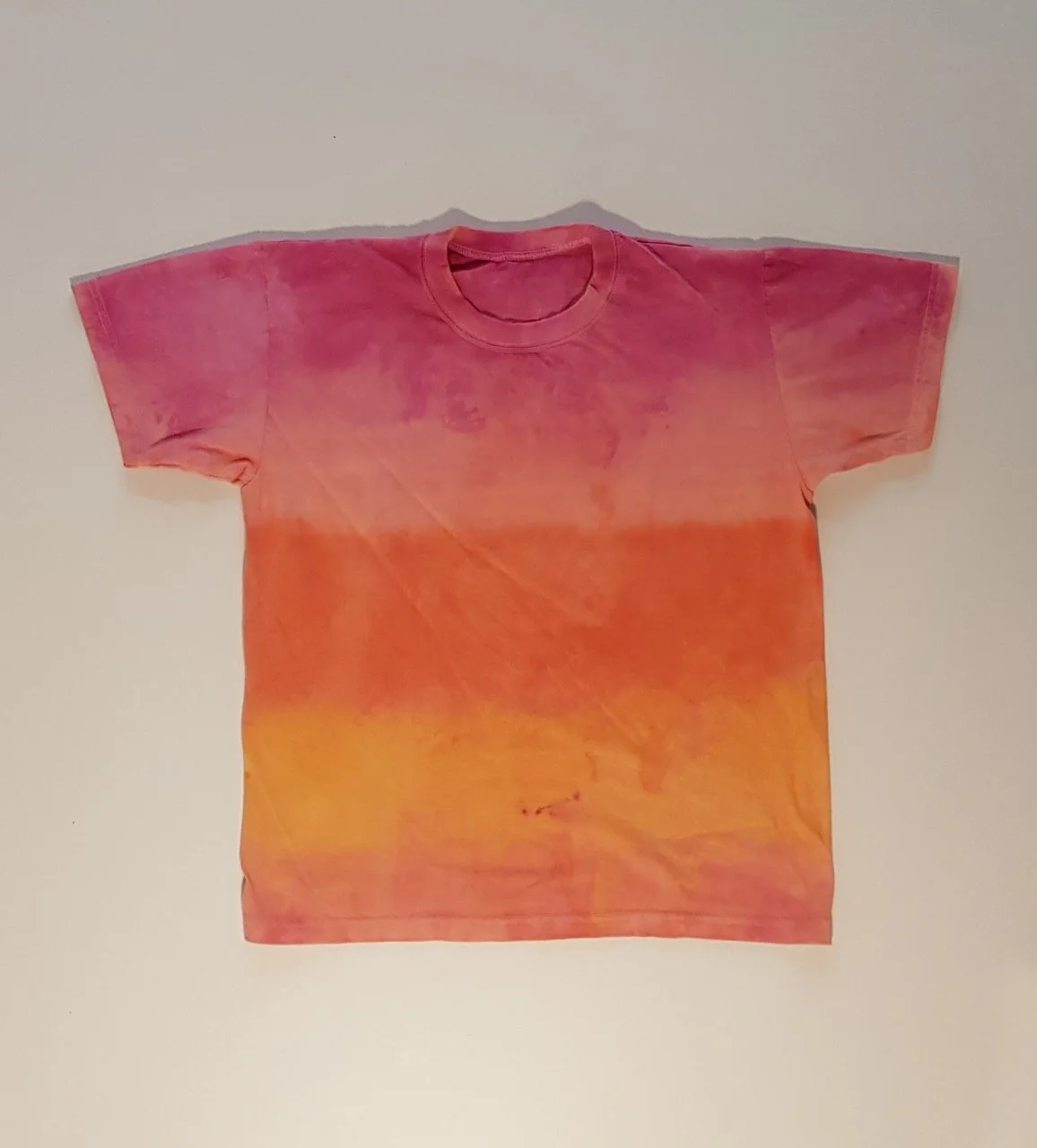A short-sleeved t-shirt with a tie-dye pattern in shades of pink, orange, and yellow. The shirt is laid flat on a plain surface.