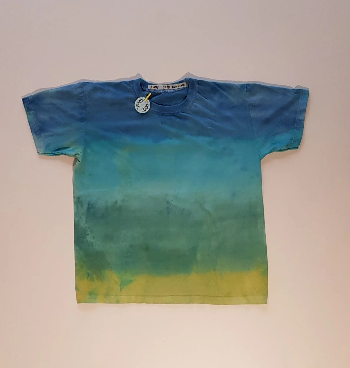 T-shirt with a tie-dye pattern in shades of blue, green, and yellow, flat on a light-colored background.