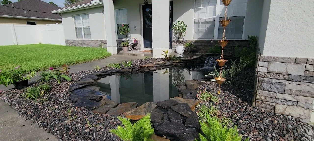 A small backyard pond with a waterfall feature, surrounded by rocks and plants, in front of a white house with a porch.