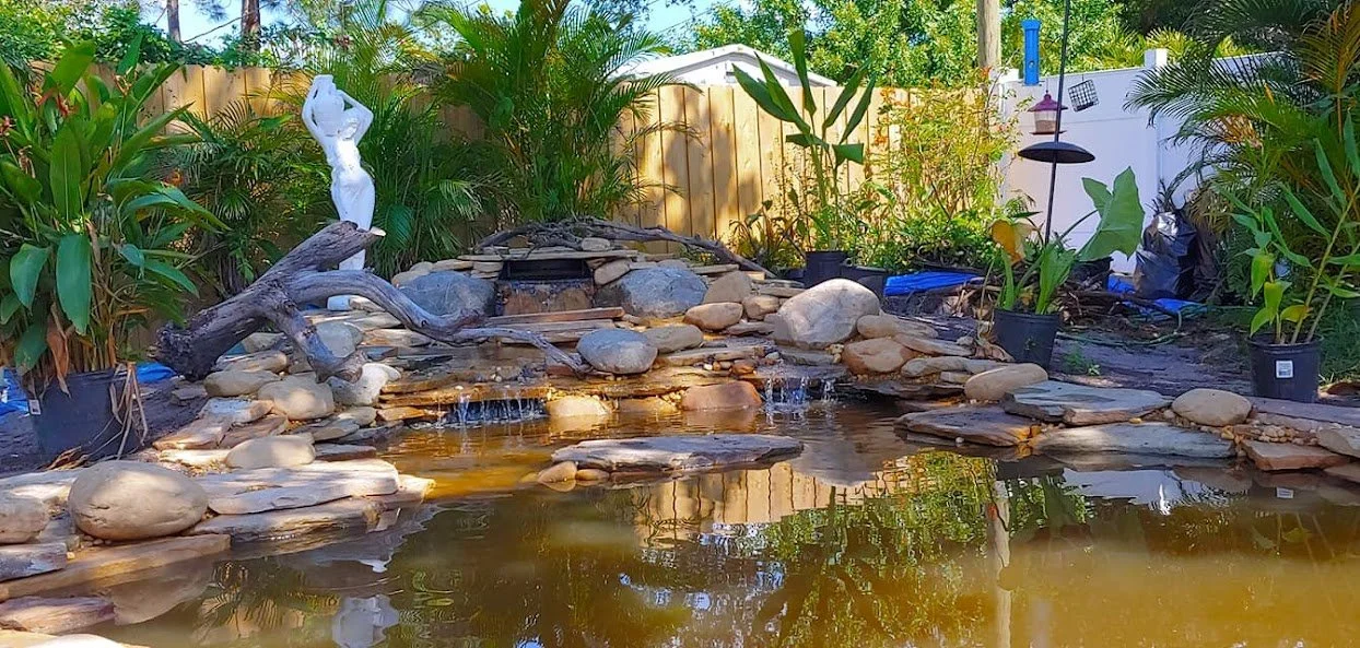 Backyard pond surrounded by rocks and plants, with a small waterfall feature, a white statue, and garden decor.