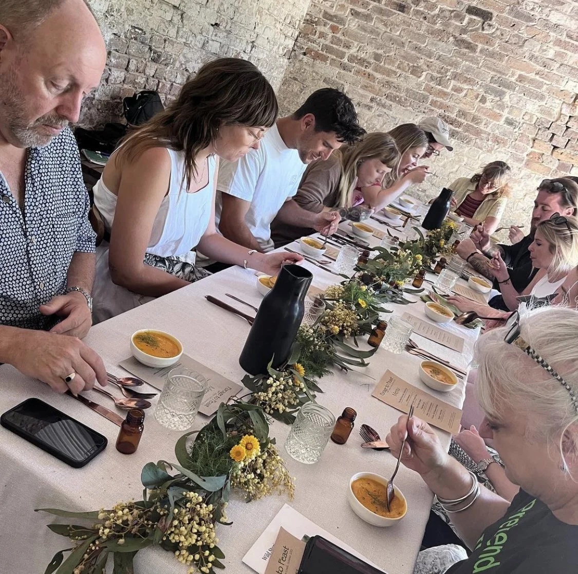 Marnee Fox A group of people sitting around a long dining table with bowls of soup, glassware, and greenery decorations on a white tablecloth. They are seated against a brick wall, focused on their meal.