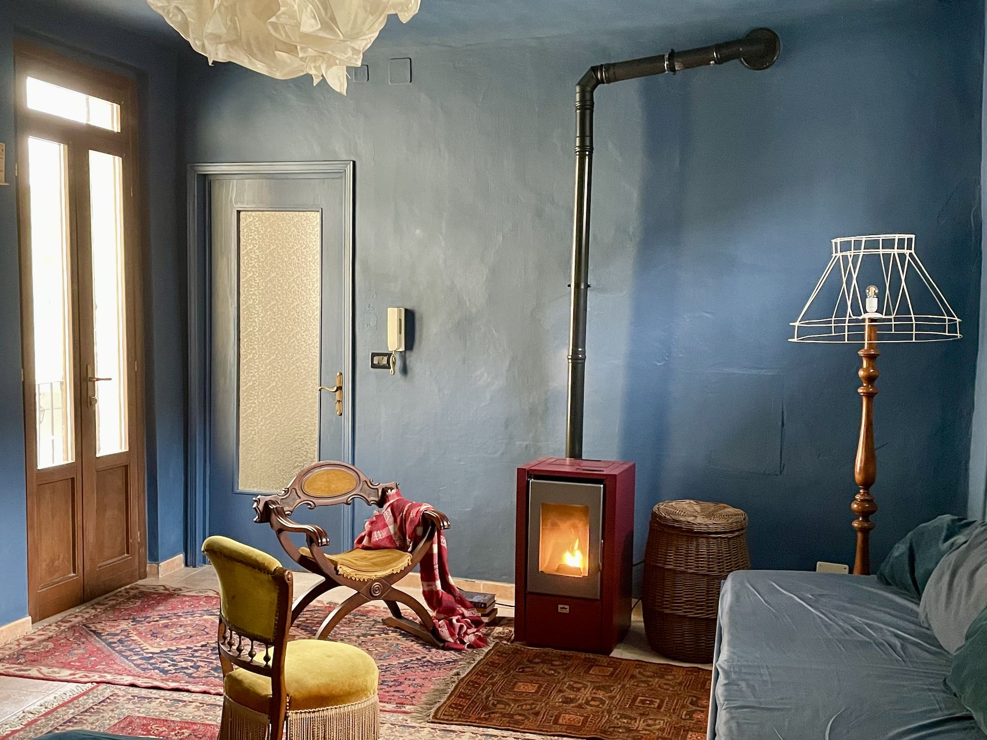 Marnee Fox Cozy living room with blue walls, featuring a wood-burning stove, wooden chair with a red throw, vintage rugs, an empty lamp stand, and wicker basket. A sofa sits against one wall with a large window and door nearby.