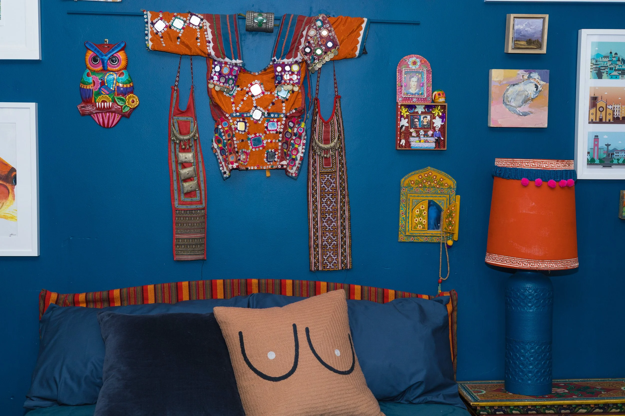 Marnee FoxEclectic room with blue walls decorated with colorful textiles and art, including a colorful owl decoration, an orange embroidered garment, framed art, and a blue and orange lamp. A bed with blue pillows and a decorative cushion sits below.