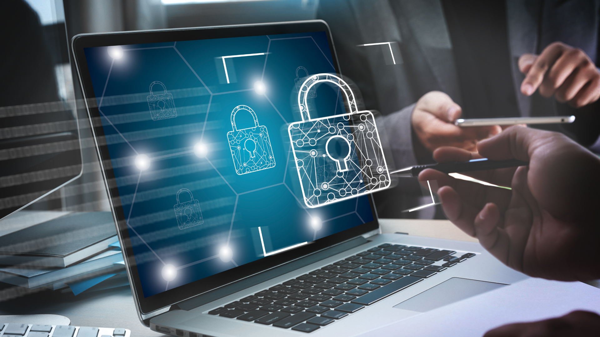 Laptop screen displaying digital padlock icons and security graphics, with a person holding a pen and a smartphone.