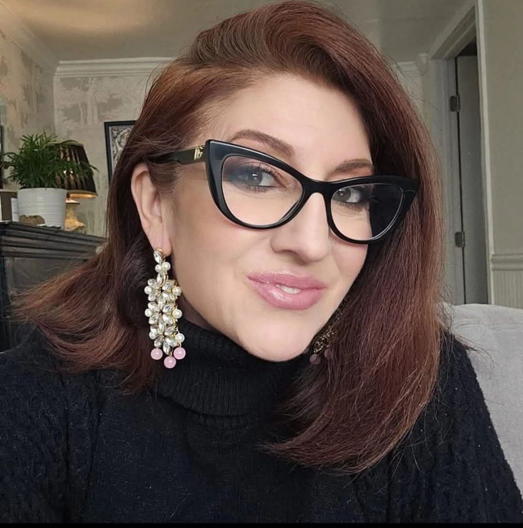 A woman with auburn hair wearing black cat-eye glasses, pearl and pink earrings, black turtleneck, smiling in a well-decorated indoor setting.