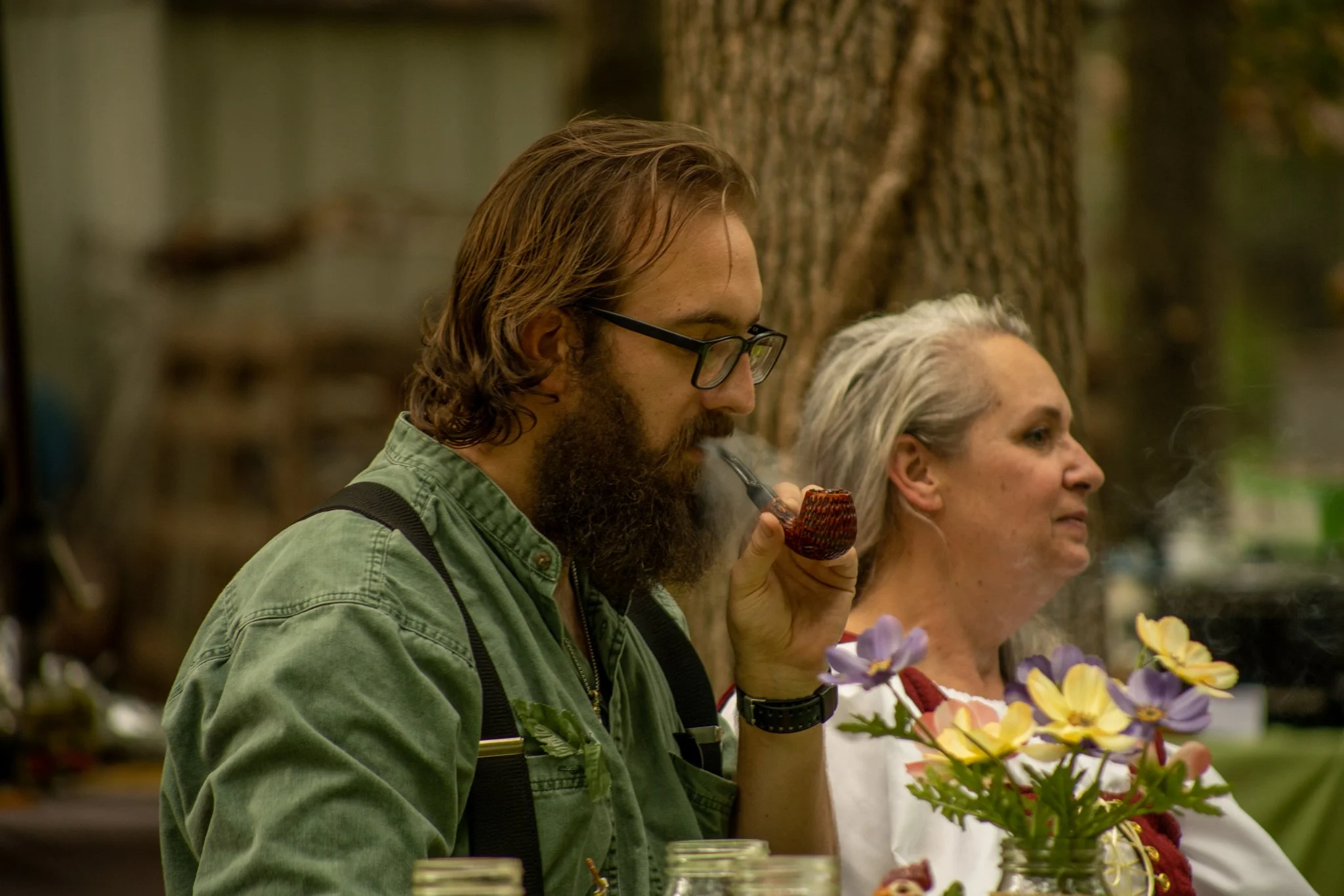 A man with glasses and a beard smoking a pipe, sitting next to an older woman with gray hair and a pained expression, in an outdoor setting with trees and flowers.