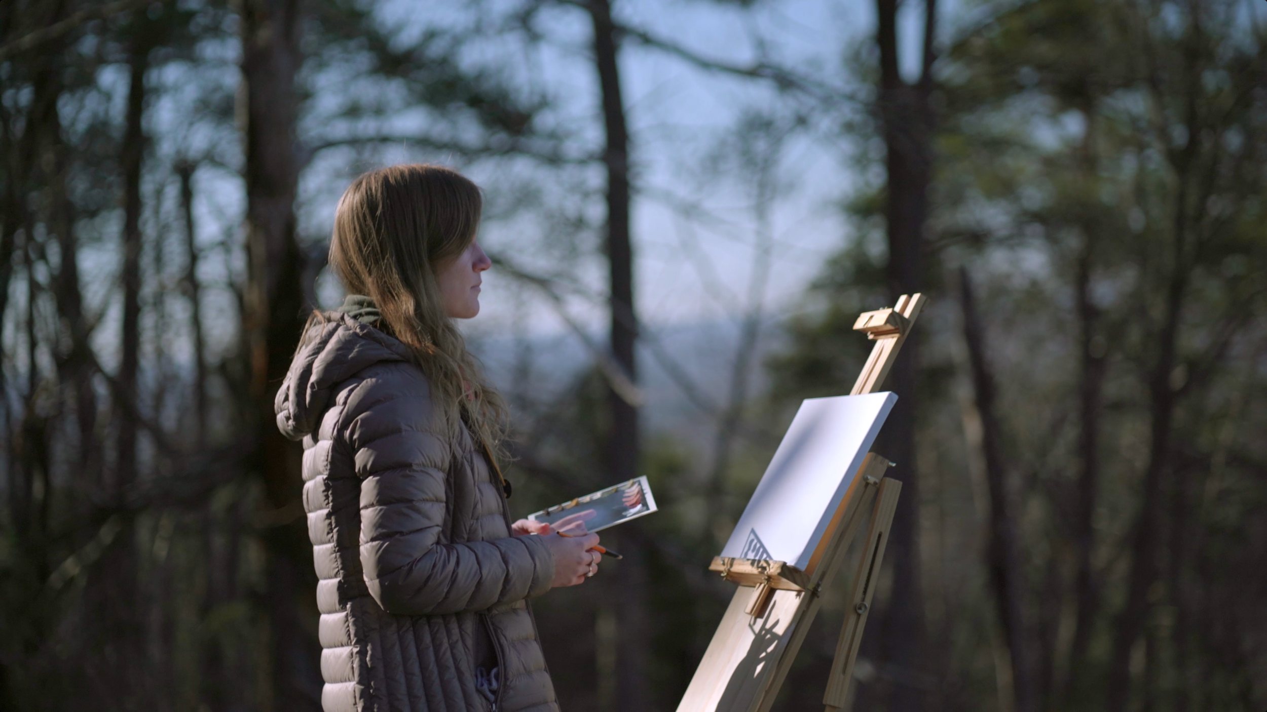 A woman in a gray puffer jacket stands outdoors in a forest, holding a palette and brush, facing a blank canvas on an easel, with trees and sky in the background.