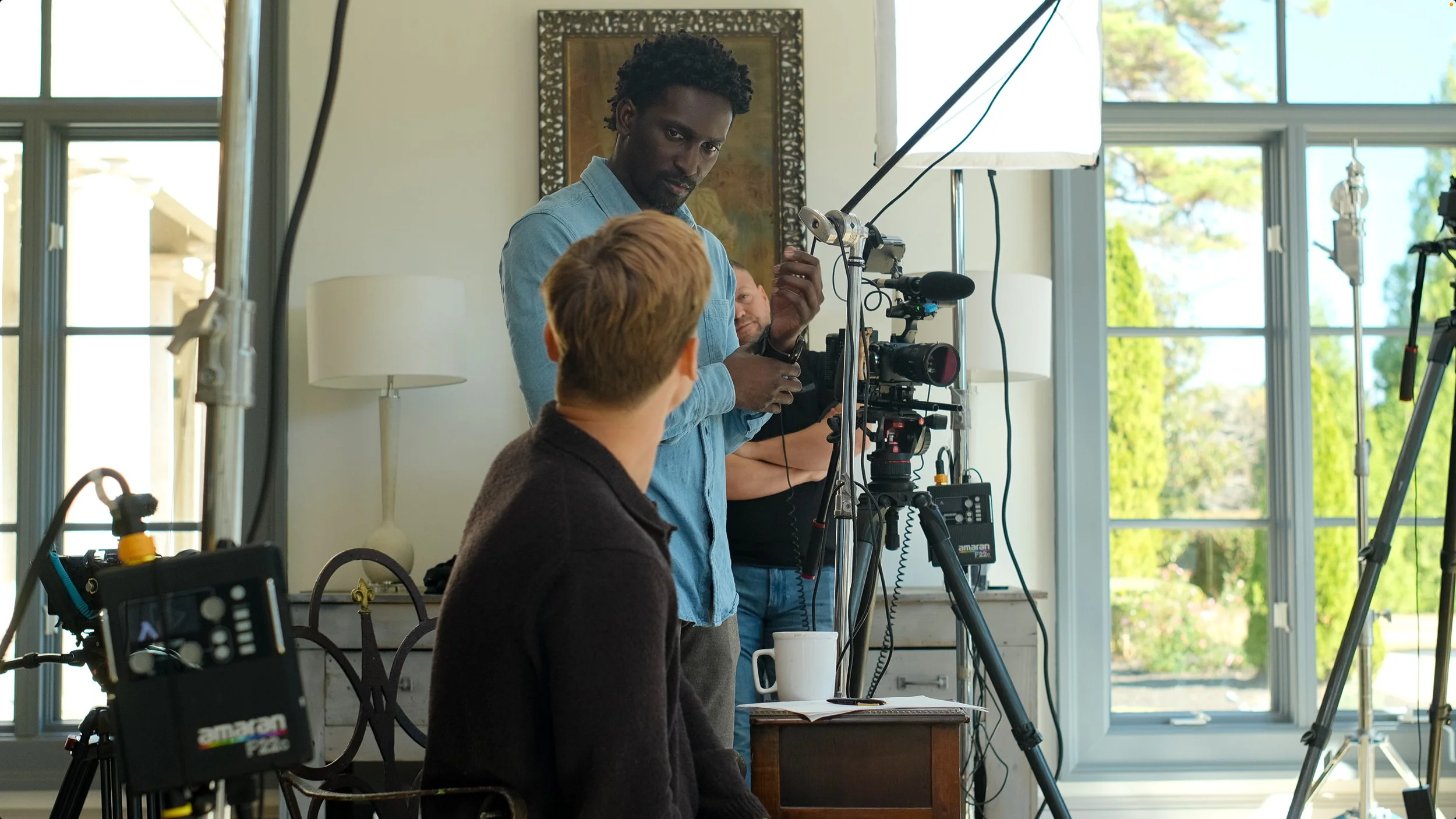 A film crew is setting up equipment for a shoot in a room with large windows, a decorative painting, and a table with a mug. One person is sitting in front of the camera, while crew members adjust cameras and lighting.