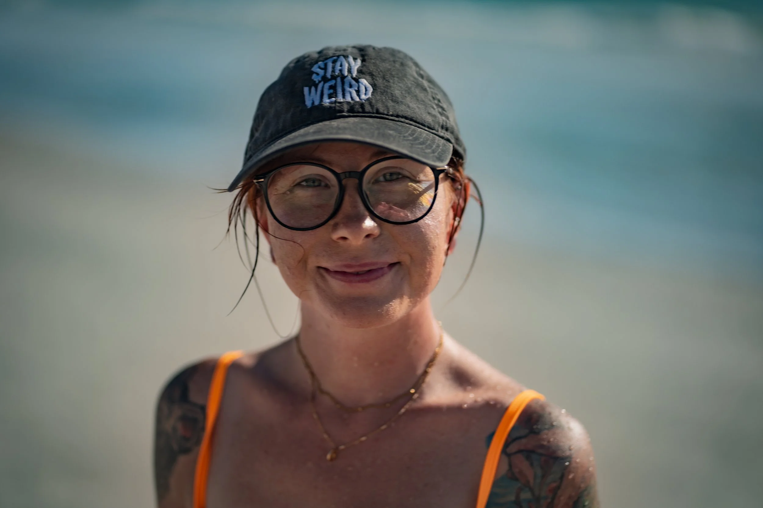 A woman with tattoos on her arms, wearing glasses, a dark baseball cap that says "Stay Weird," and an orange swimsuit, smiling on a beach with water in the background.