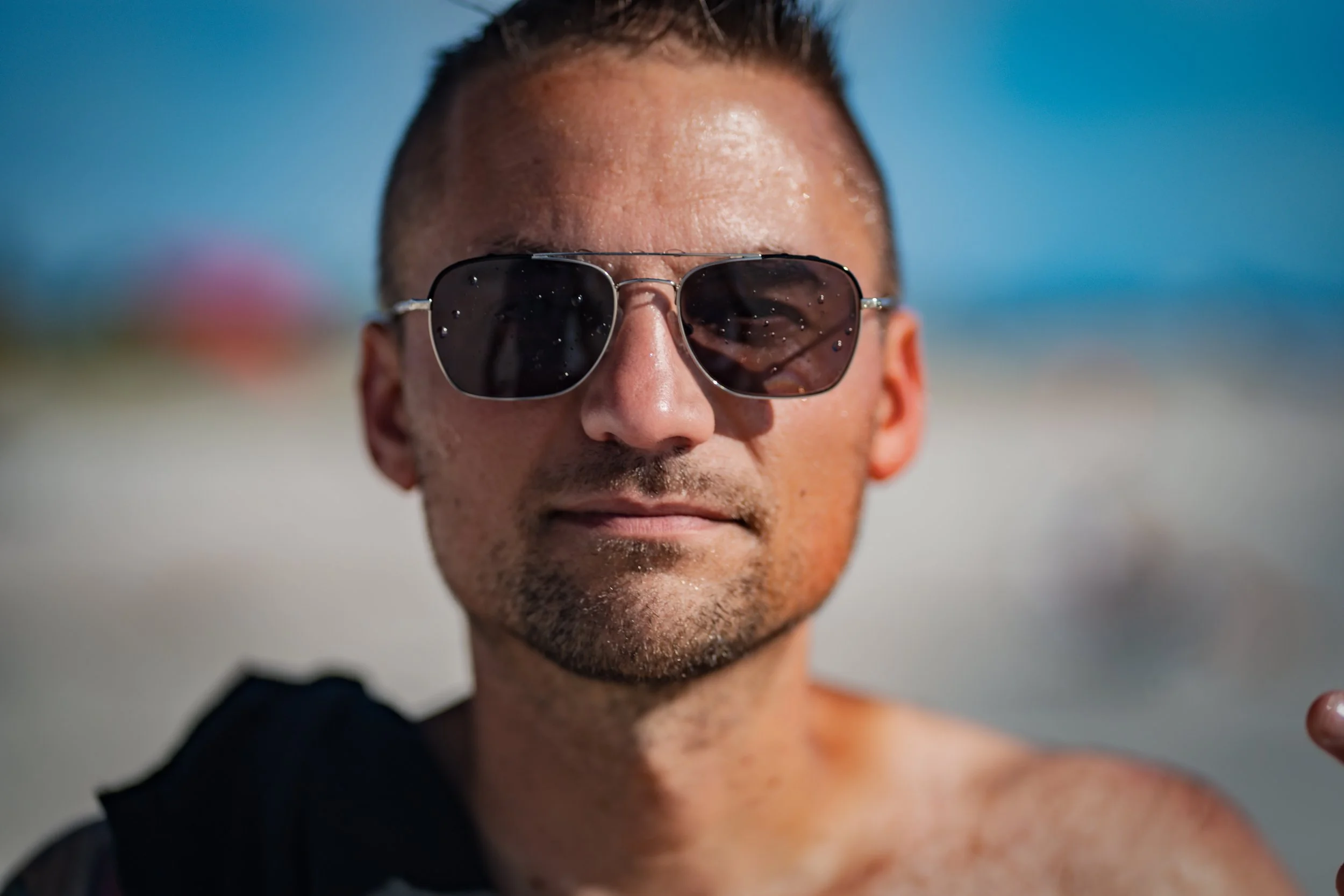 A man wearing sunglasses at the beach with water droplets on his face, a blurred beach background, and a black towel over his shoulder.