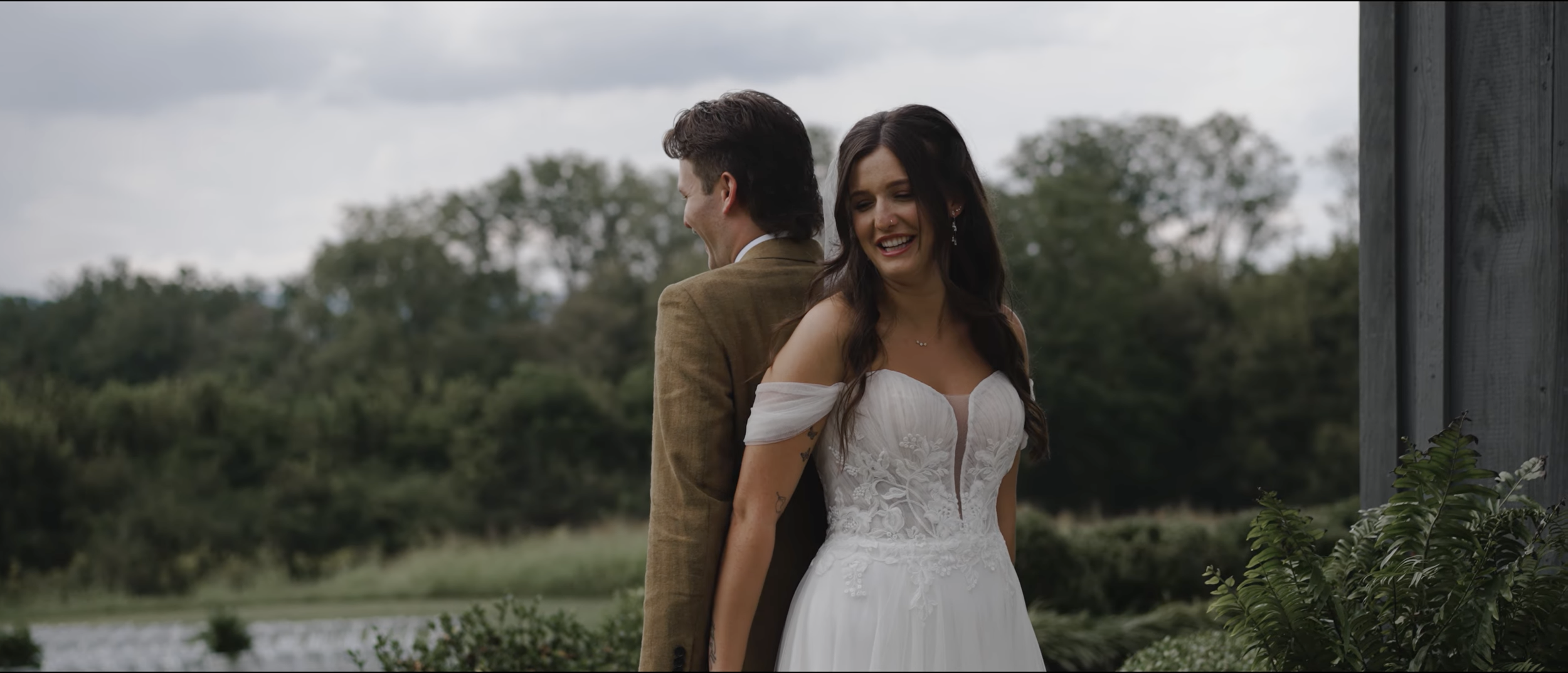 A couple, dressed in wedding attire, stand back to back outdoors with trees and a cloudy sky in the background. The woman is smiling and looking away, wearing a white off-shoulder wedding dress with lace, and the man is looking to the side, wearing a brown suit jacket.