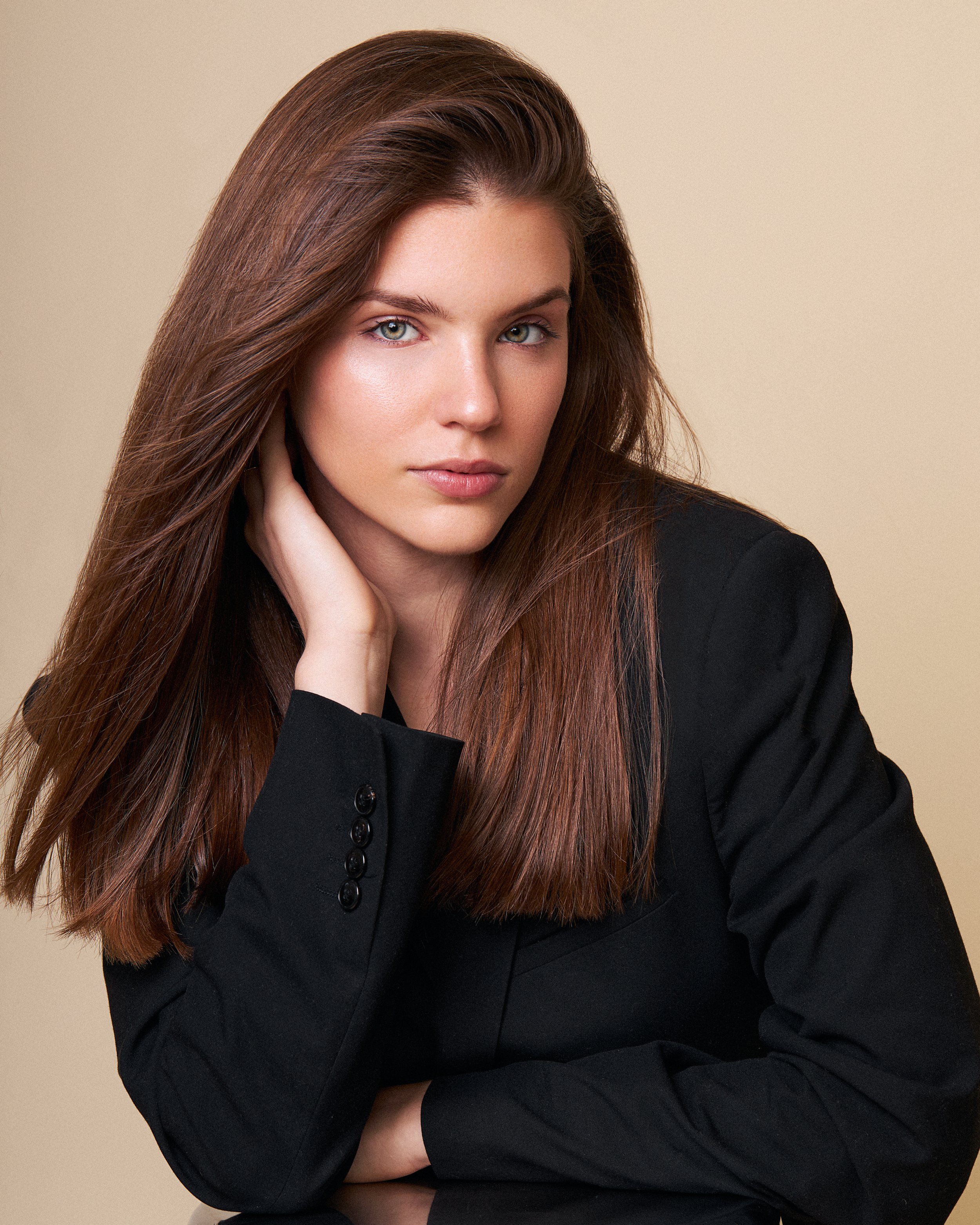 Retrato de una mujer con cabello largo y castaño, usando saco negro, posando con la mano en la cara, fondo beige.