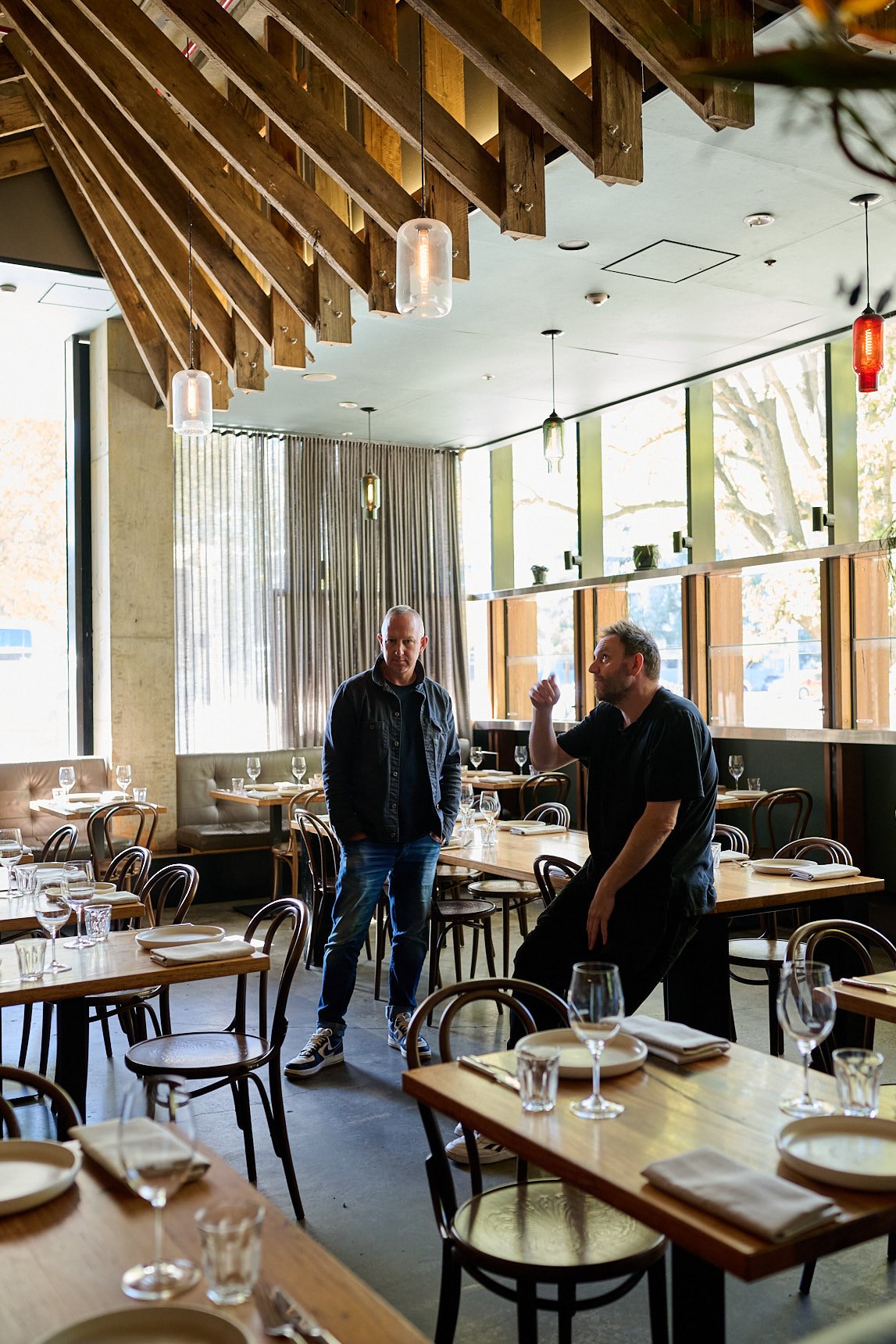 Two men in a modern, well-lit restaurant with wooden tables and chairs, set with plates, glasses, and napkins, engaged in conversation.