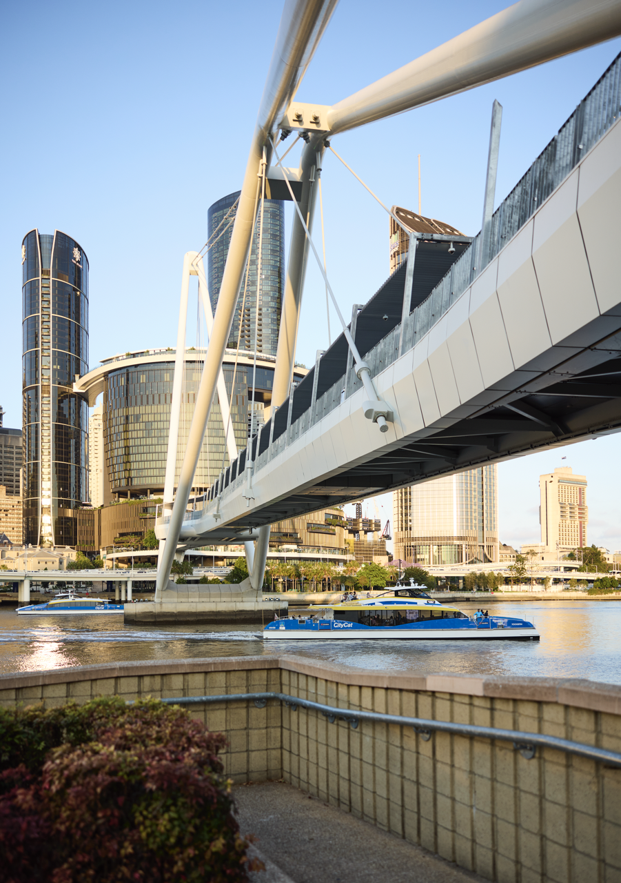 A cityscape view with modern skyscrapers and a footbridge over a river, with a blue and yellow CityCat passenger ferry on the water.