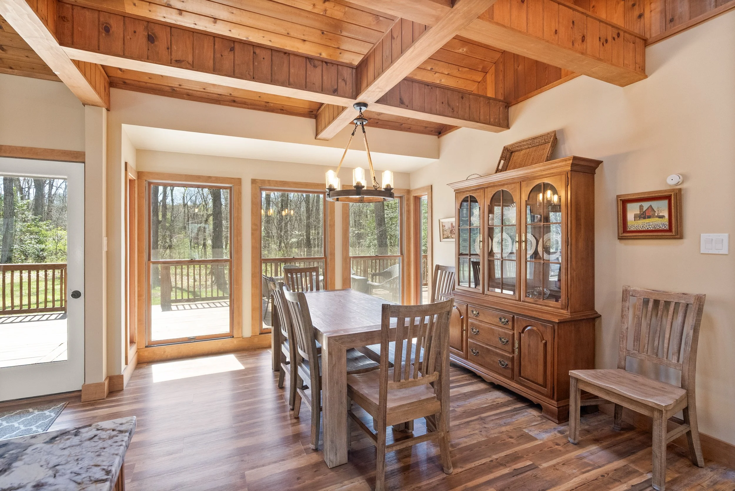 Wooden dining room with a table, six chairs, a china cabinet, large windows, and a wooden ceiling with beams.