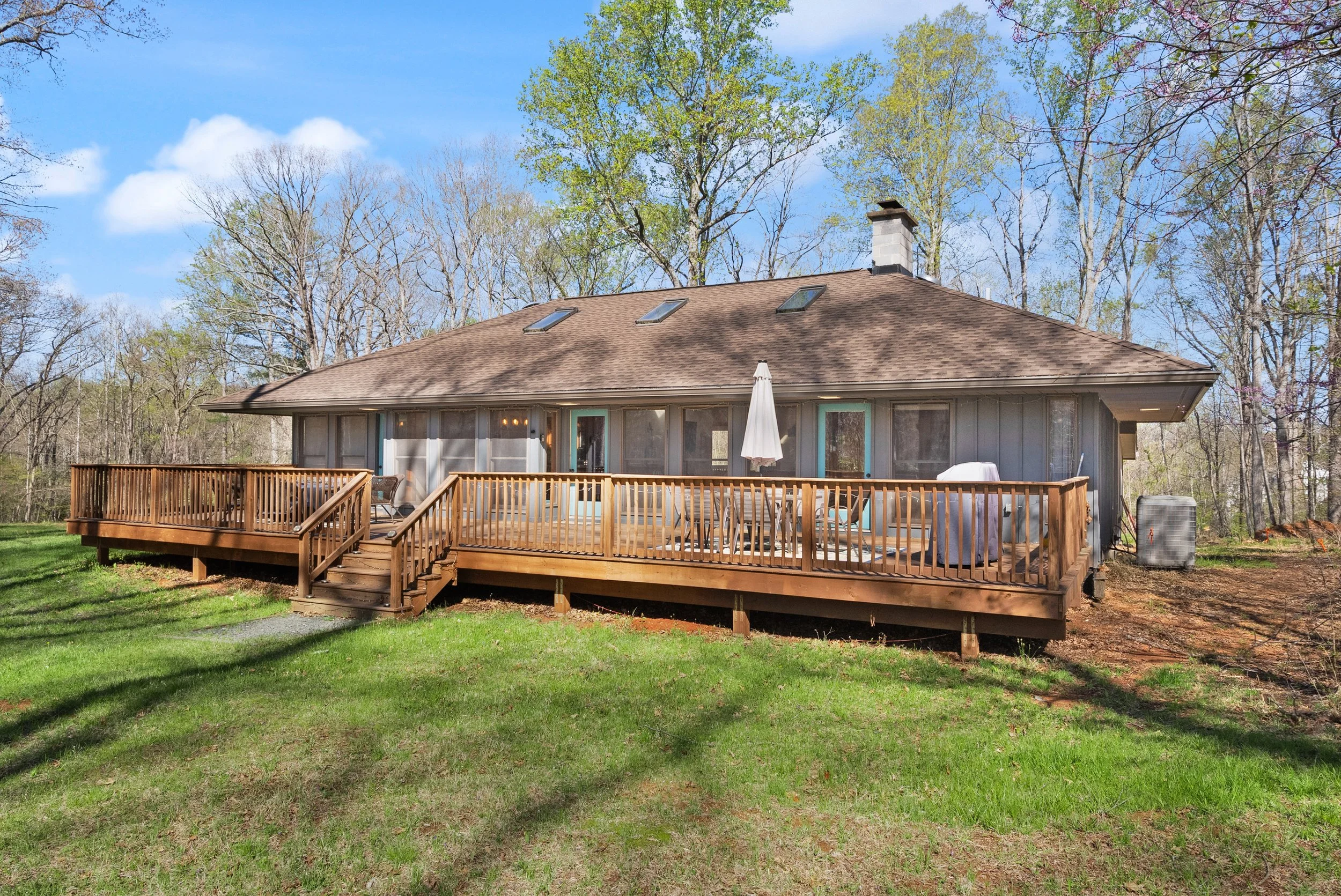 A house with a large wooden deck, surrounded by trees and green grass, under a blue sky with scattered clouds.