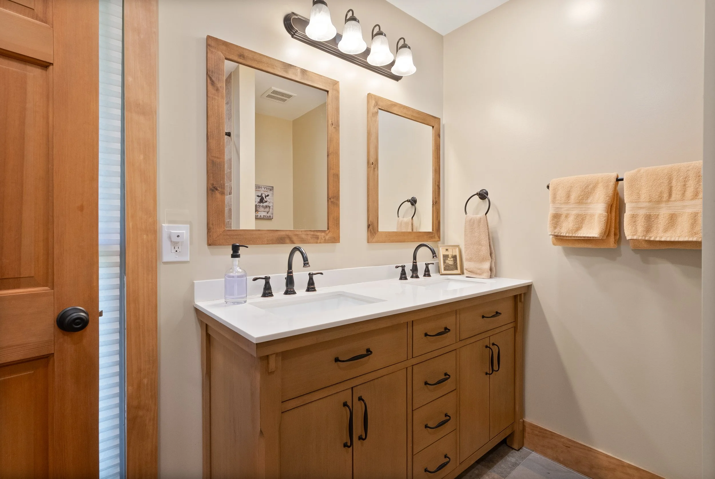 Bathroom with double sink vanity, wooden framed mirrors, beige towels, and black fixtures.