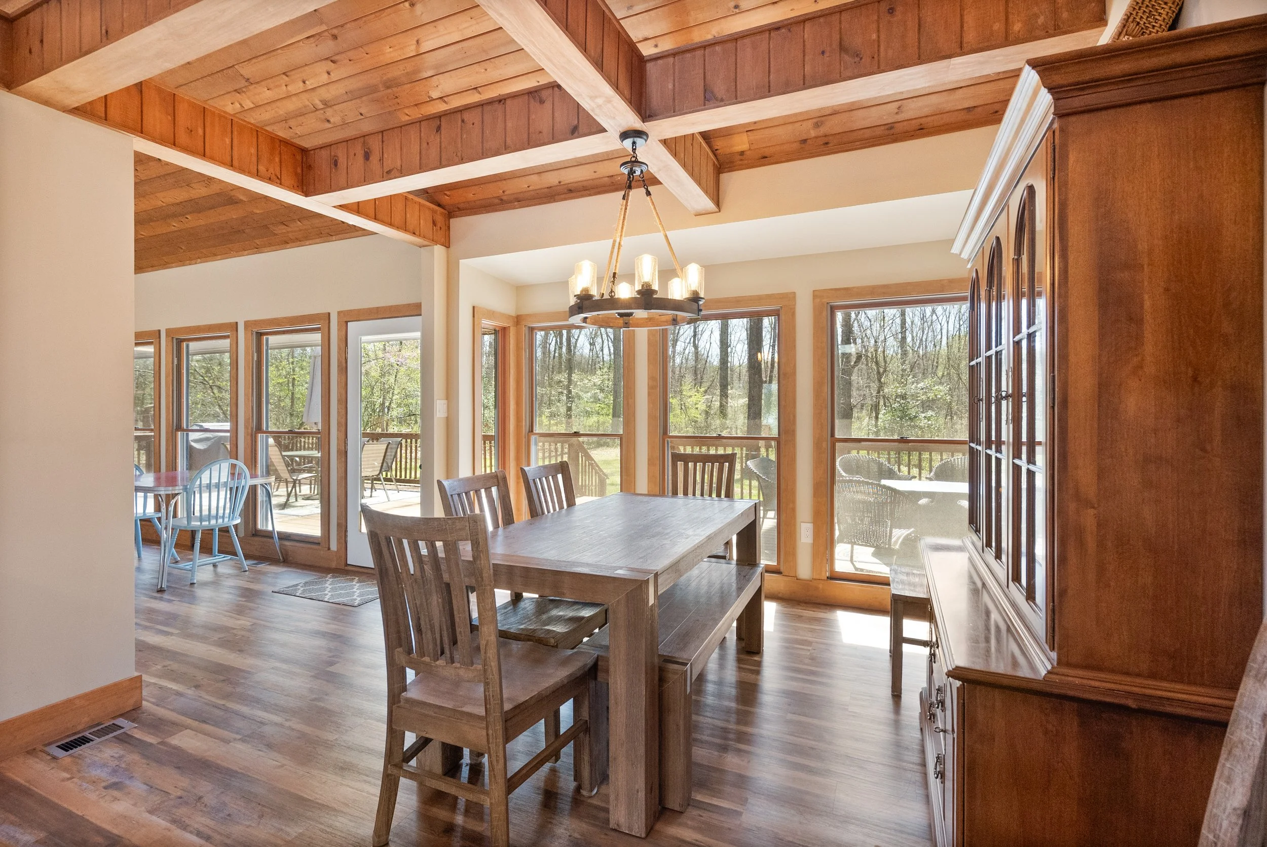 Dining area with wooden table and chairs, large windows showing outdoor patio and trees, wooden ceiling with exposed beams, chandelier hanging above the table, and a large wooden cabinet.