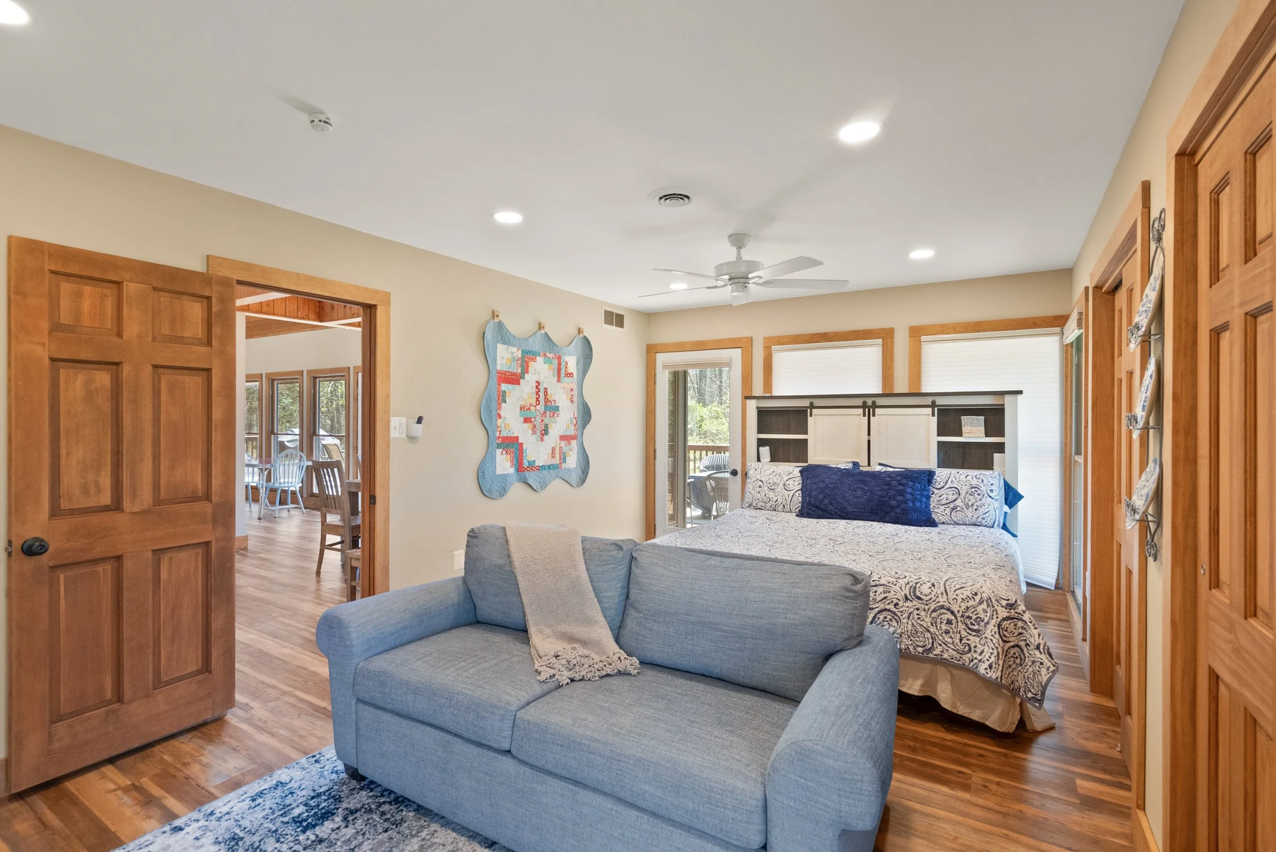 Bedroom with wooden trim and a sliding glass door leading to patio, featuring a bed with patterned bedding, a blue pillow, a wooden headboard, a light blue sofa with a beige throw blanket, and wall art, with hardwood floors and recessed lighting.