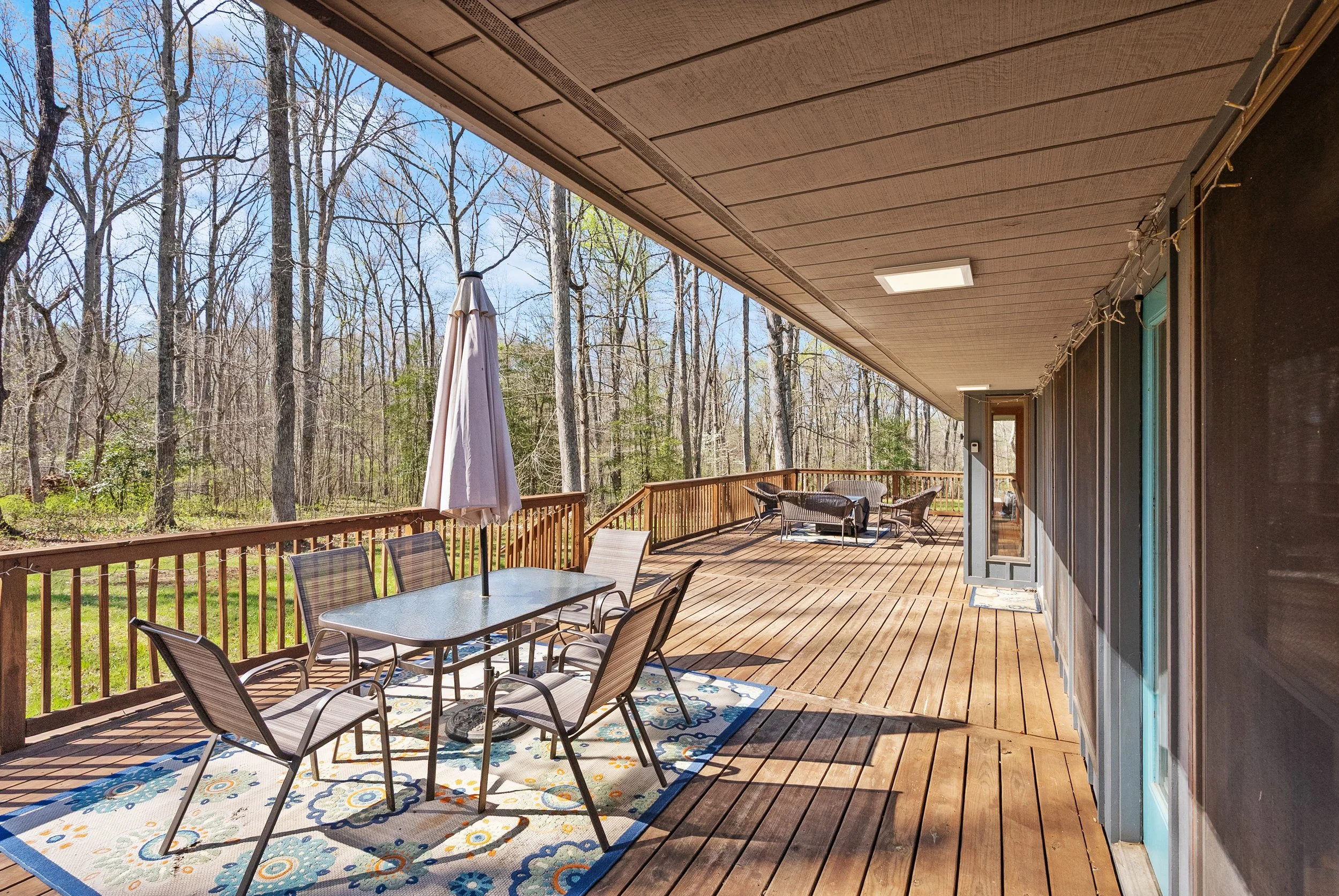 Large wooden deck with outdoor furniture, including a table with six chairs, a patio umbrella, and a seating area with a sofa and chairs, surrounded by leafless trees under a clear sky.