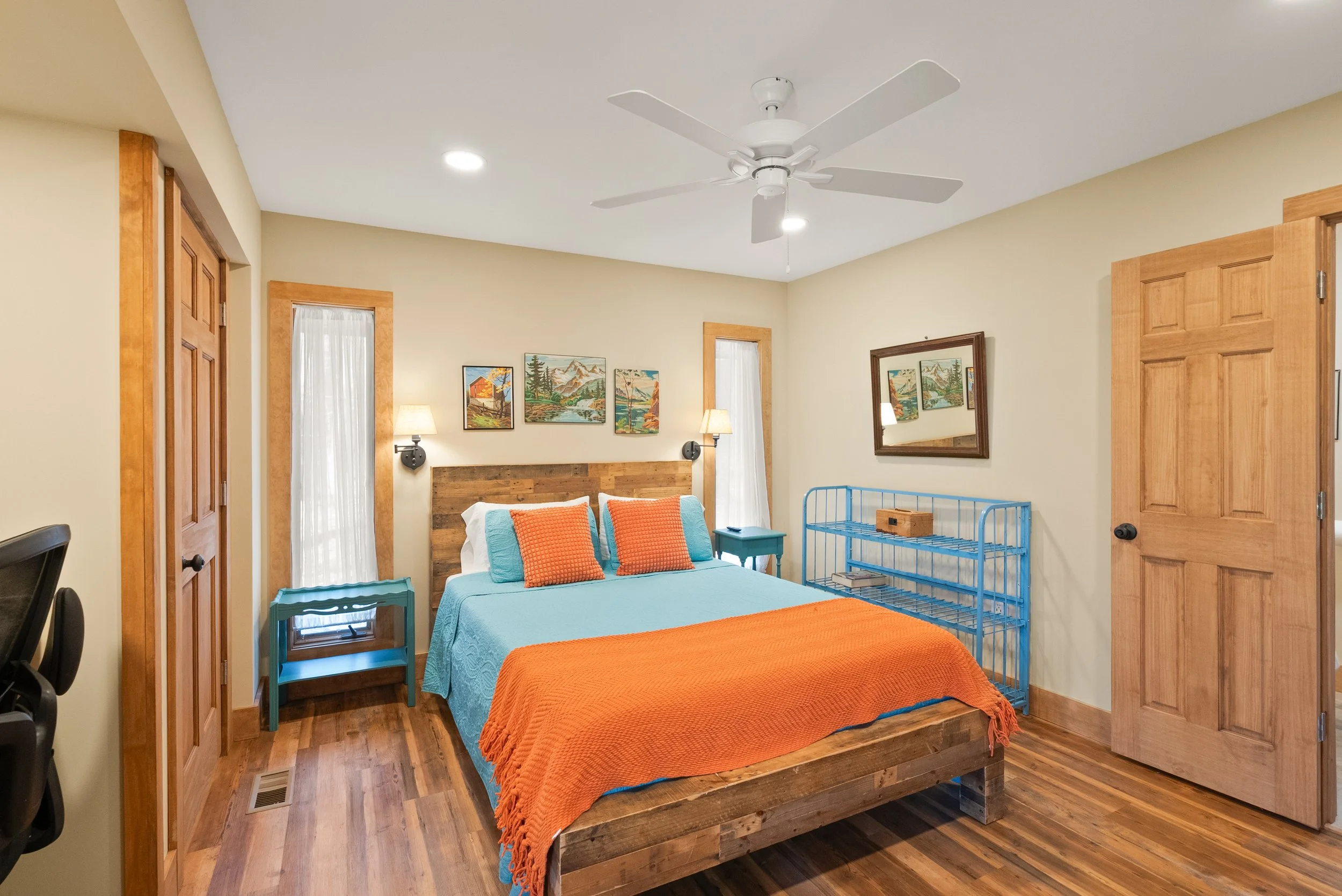 A bedroom with a wooden bed frame, blue bedspread, orange blanket, and matching orange pillows. There are two nightstands, each with a lamp, and framed artwork above the bed. The room features wooden window frames, a wooden door, and a blue metal shelf. The floor is wood, and there is a ceiling fan.