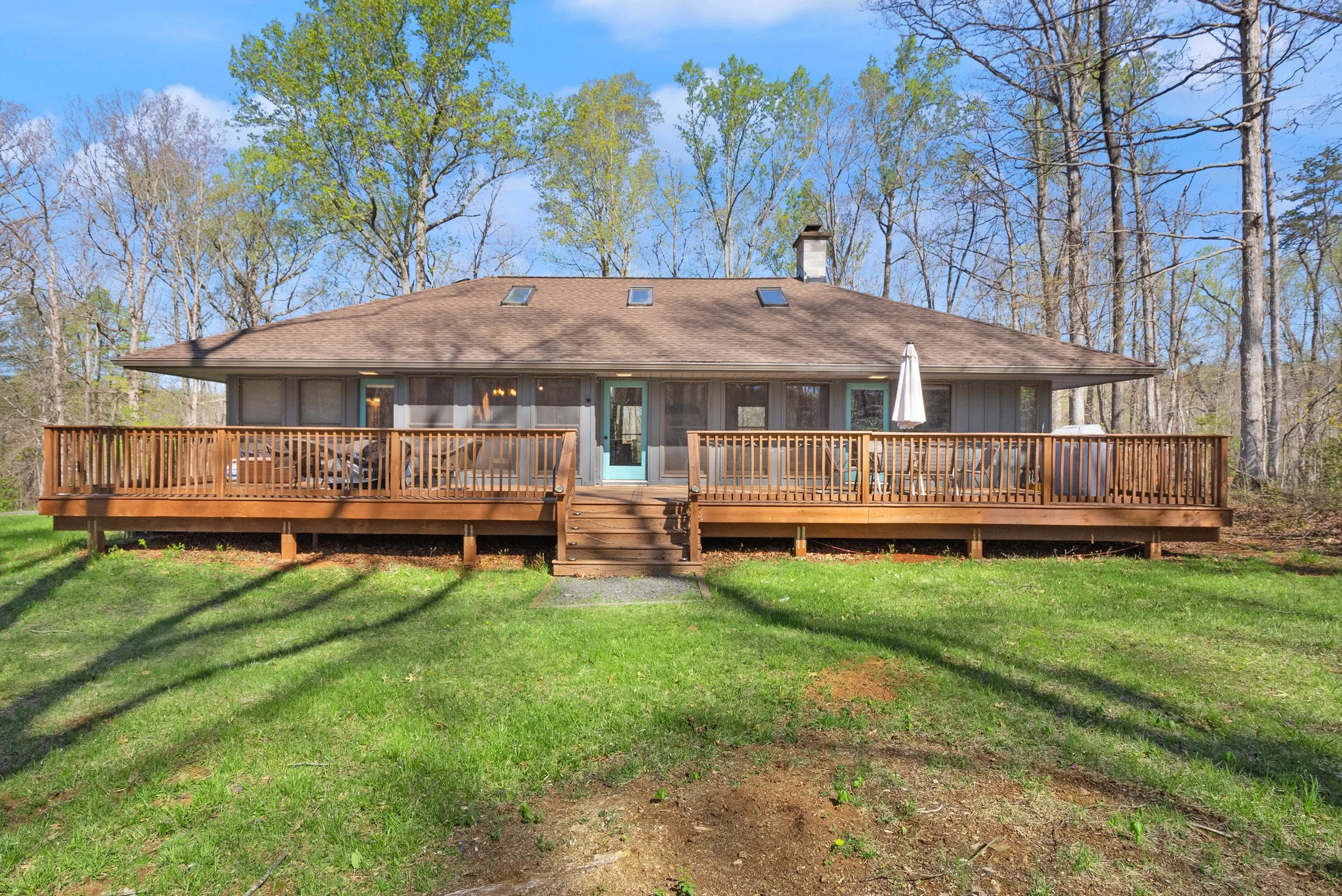Backyard view of a house with a large wooden deck, sliding glass door, and patio furniture, surrounded by trees and a green lawn.