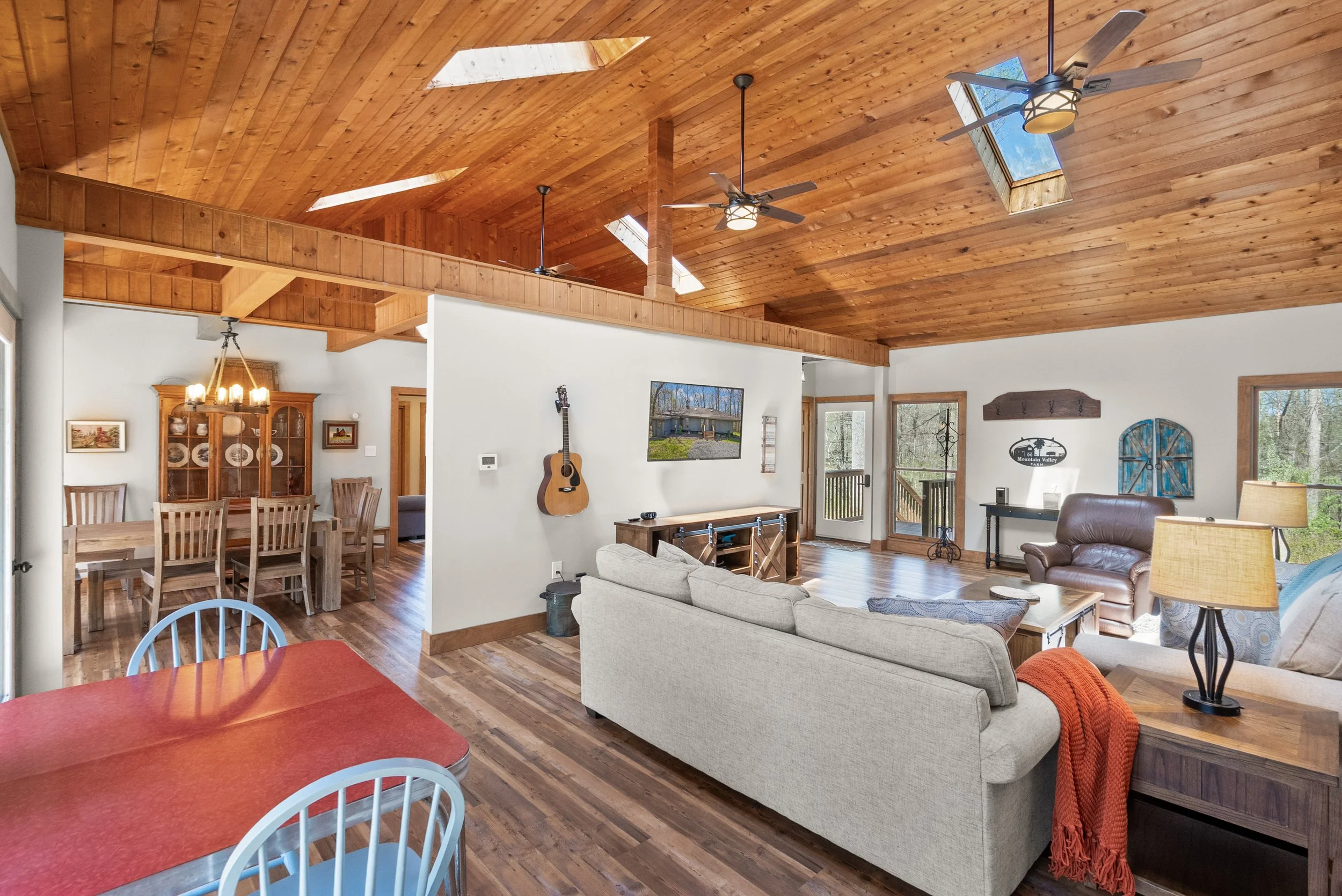 Living room with wooden vaulted ceiling, skylights, ceiling fans, a television, a beige sofa, a brown armchair, and large windows showing a wooded area outside.