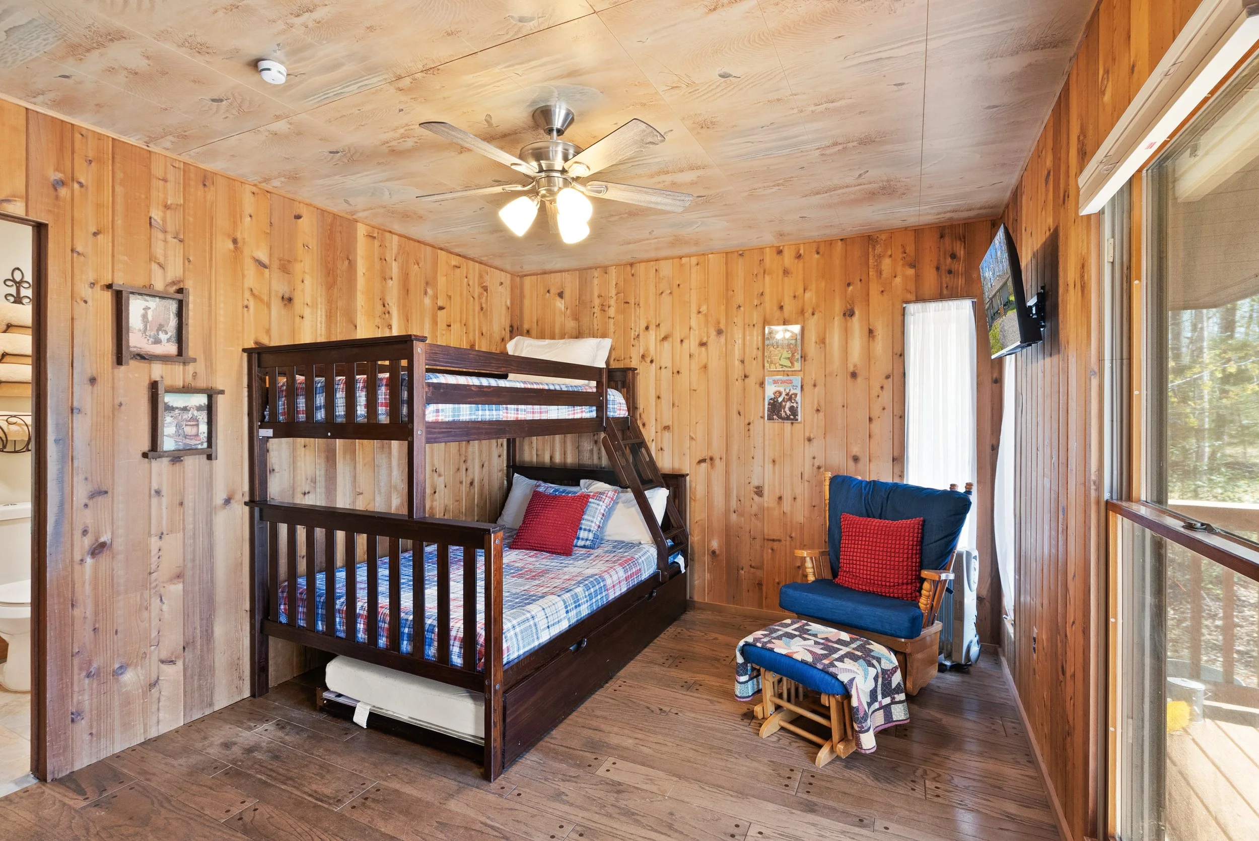 Cozy room with wooden walls and ceiling, featuring a bunk bed with plaid bedding, a rocking chair with a red pillow, a window with vertical blinds, and a small television on the wall.