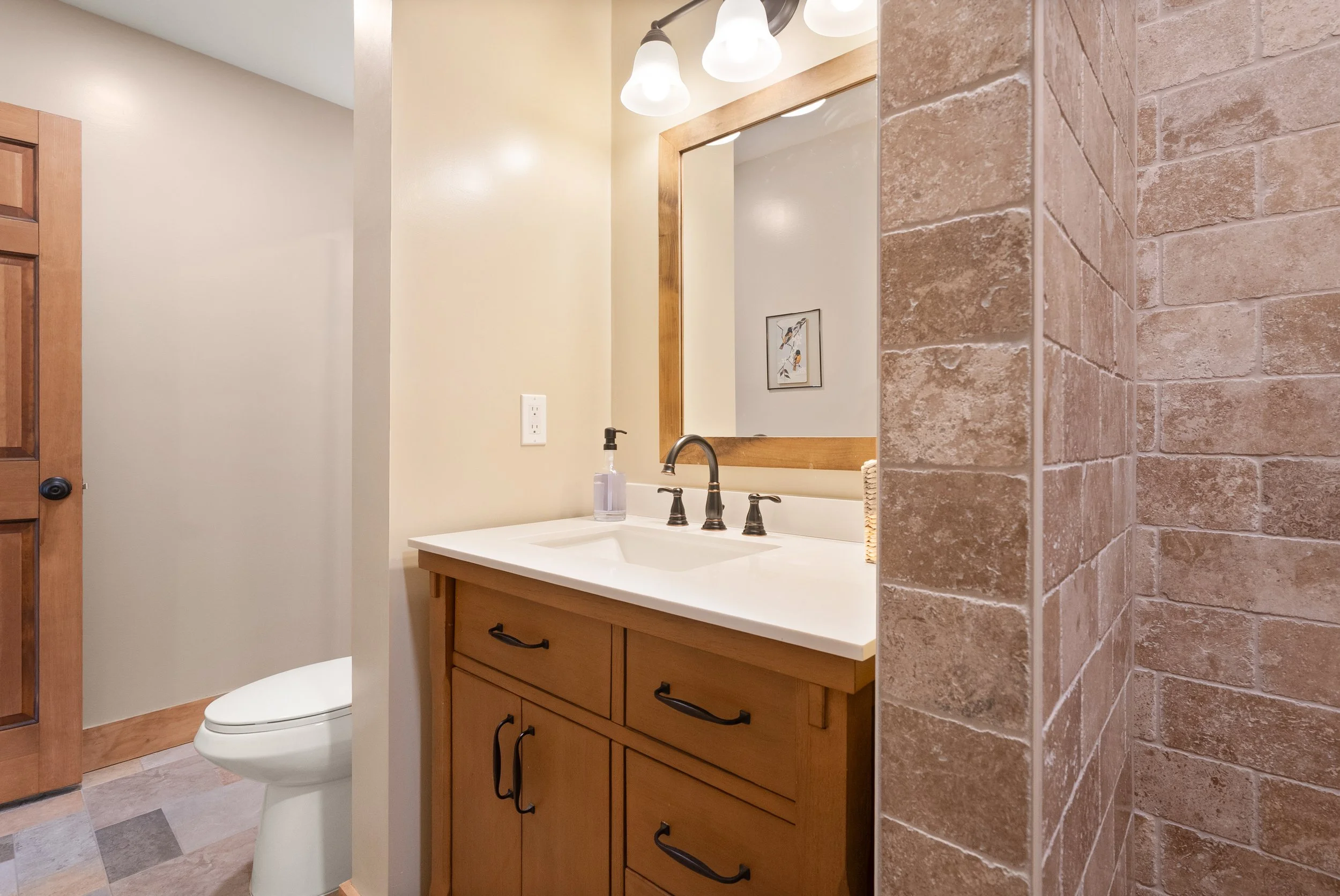 Bathroom with a wooden vanity, white countertop, and mirror above the sink. There are three light fixtures above the mirror. Part of a toilet and a brown tiled wall are visible.
