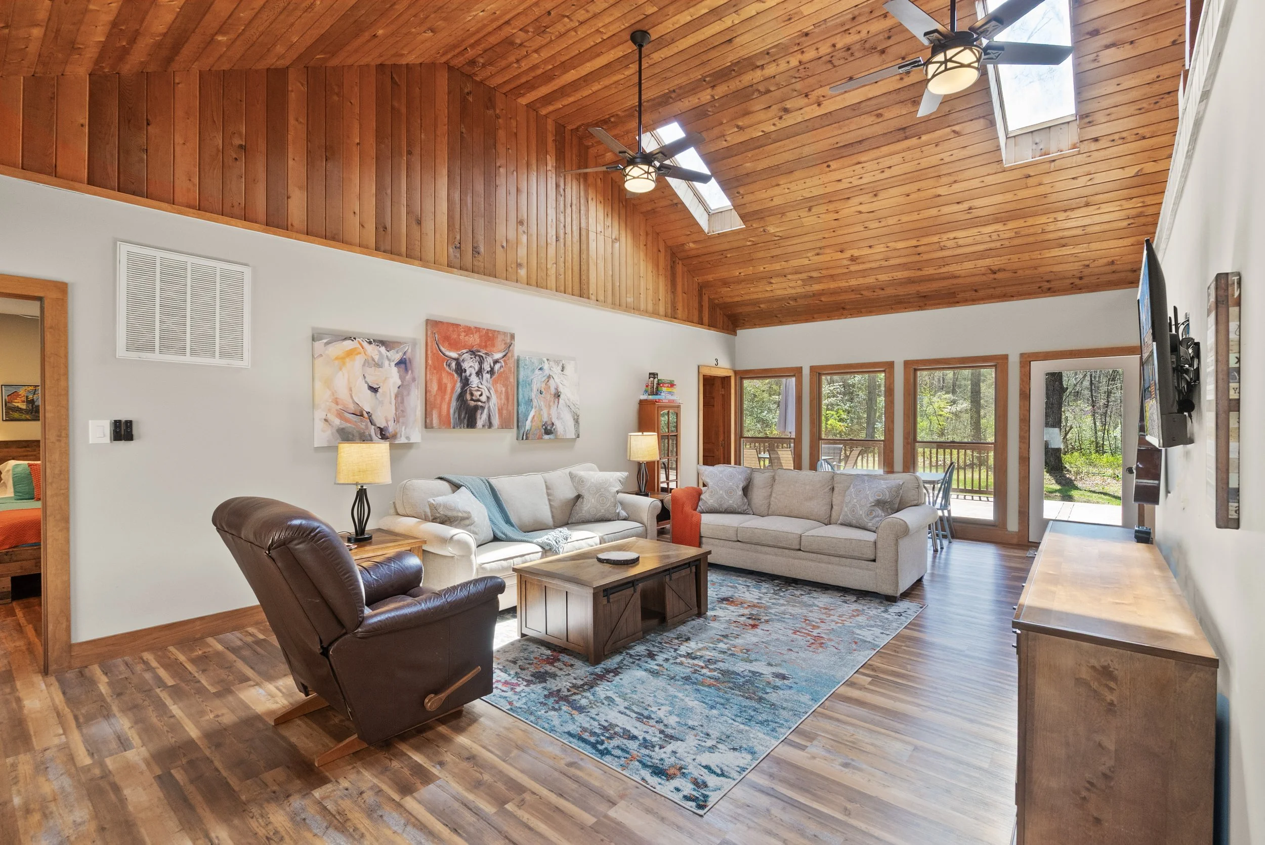 Spacious living room with wooden vaulted ceiling, skylights, and large windows, furnished with sofas, an armchair, a wooden coffee table, and a colorful area rug, decorated with animal-themed artwork.
