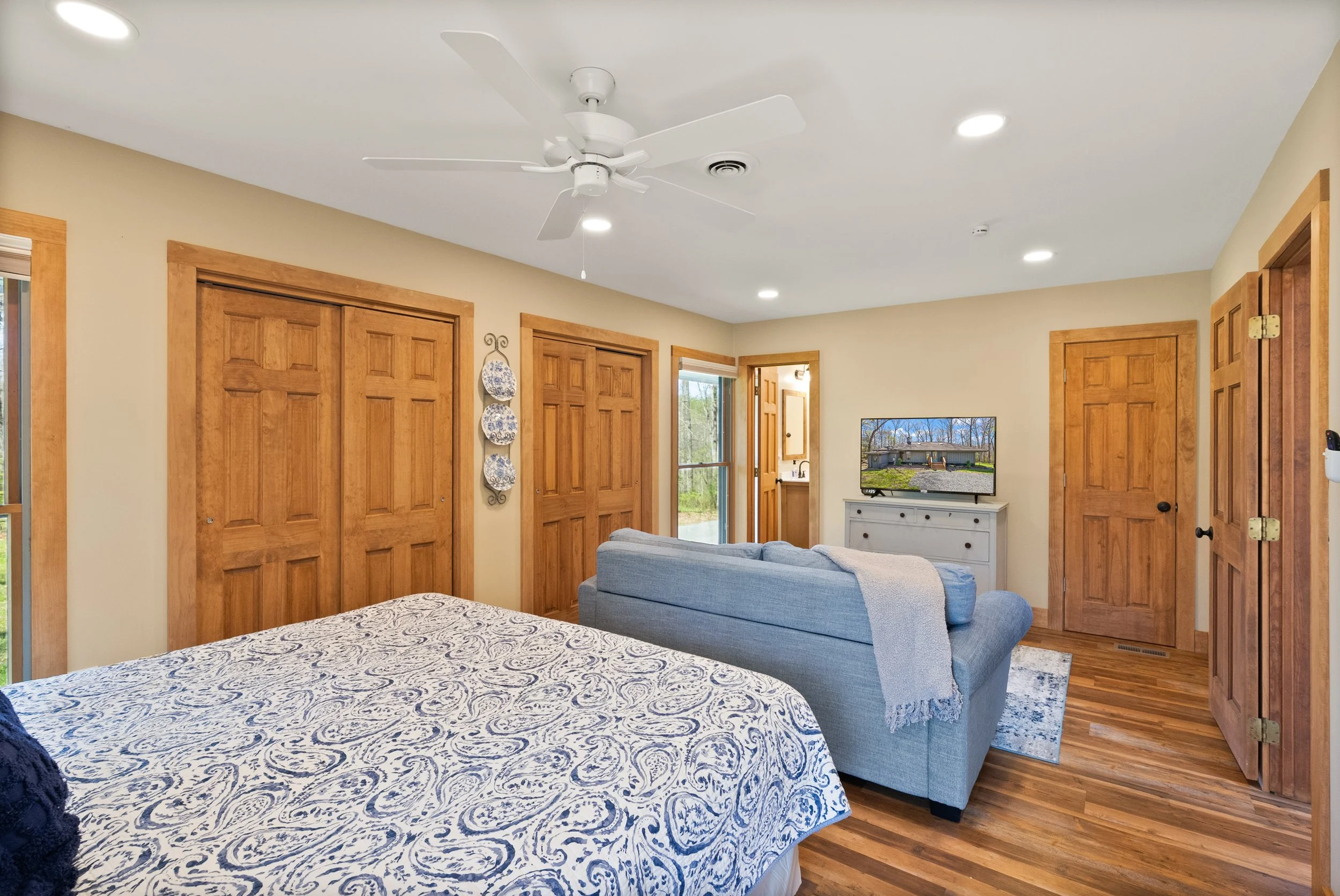 A cozy bedroom with a bed, a blue sofa, a TV on a white dresser, wooden closet doors, and a ceiling fan.