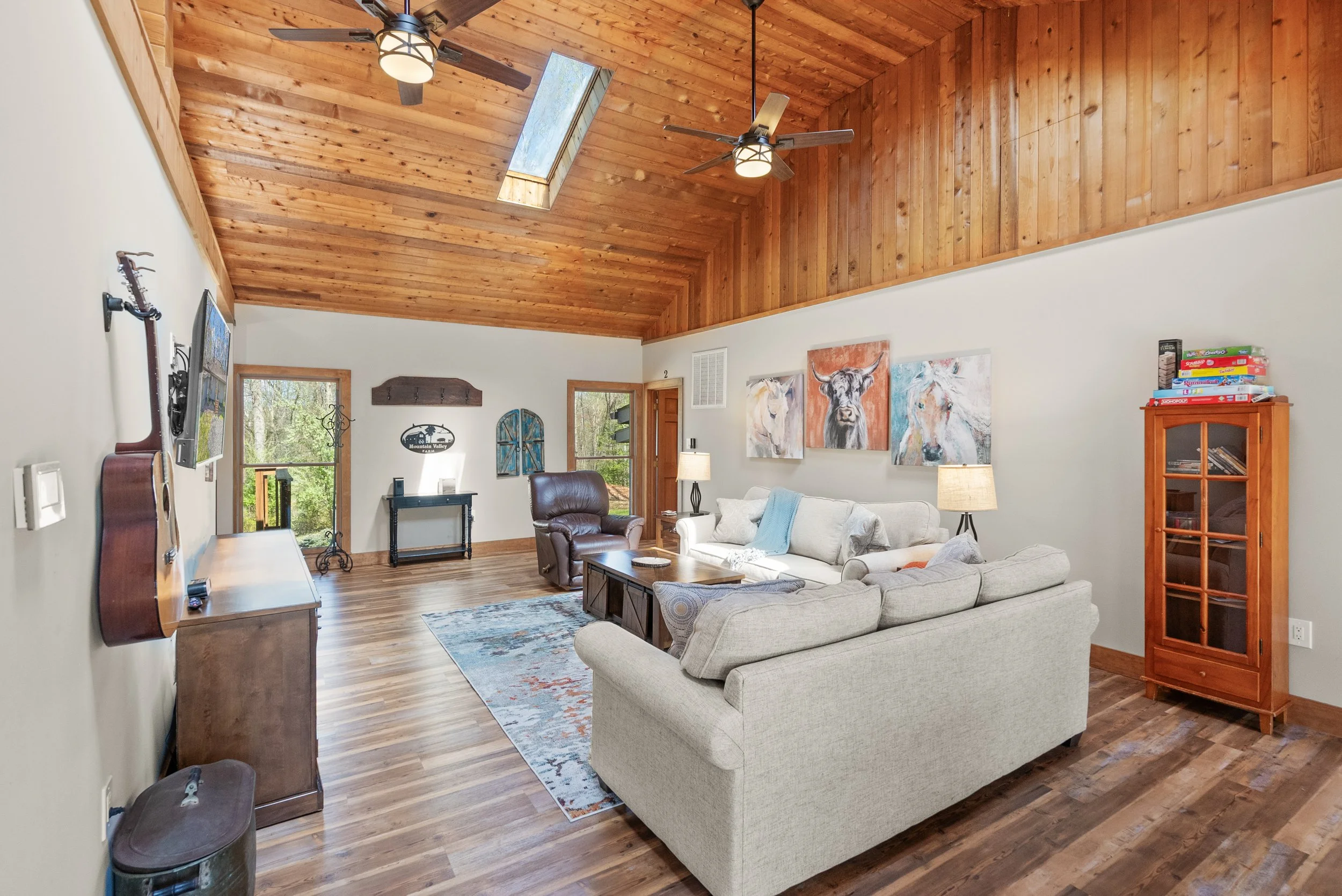 Living room with a sloped wooden ceiling, skylight, ceiling fans, and hardwood floors, furnished with a beige sectional sofa, a brown leather armchair, and various artwork on the walls.