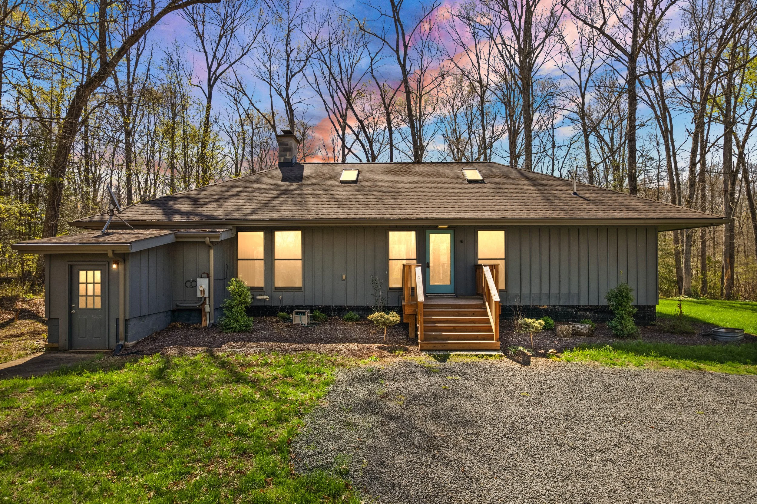 Single-story house with gray siding, front wooden steps, three large windows, wooden porch, surrounded by leafless trees and green grass, sunset sky with pink and purple clouds.