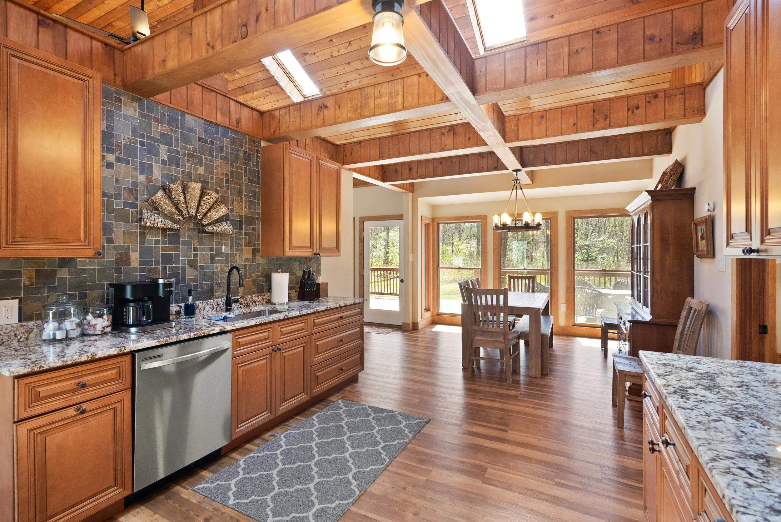 Open-concept kitchen and dining area with wooden cabinets, granite countertops, a tile backsplash, and large windows overlooking a backyard.