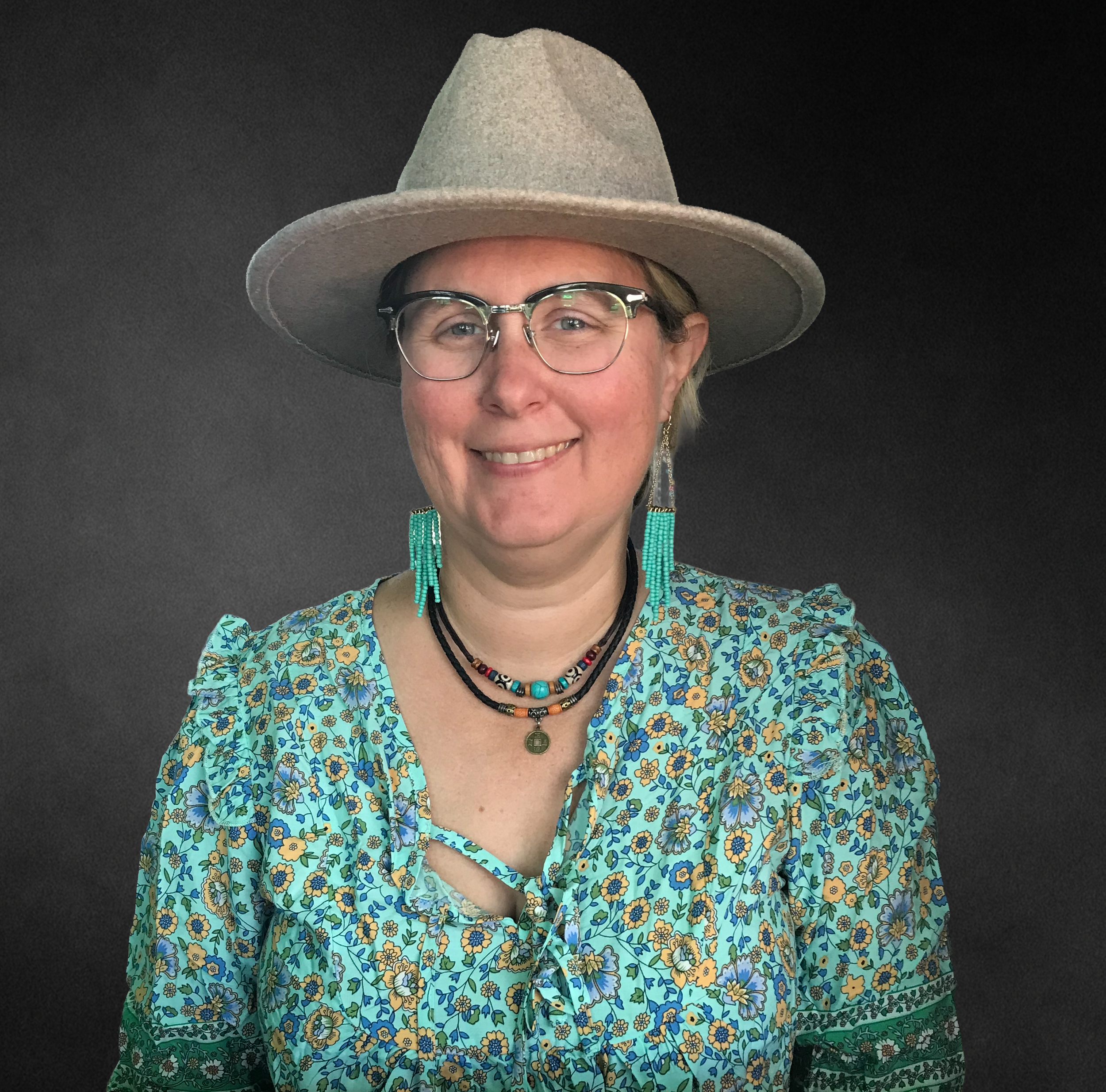 A woman wearing a beige wide-brimmed hat, glasses, turquoise beaded earrings, a black beaded choker, and a floral shirt with blue, yellow, and green colors, standing against a dark background.