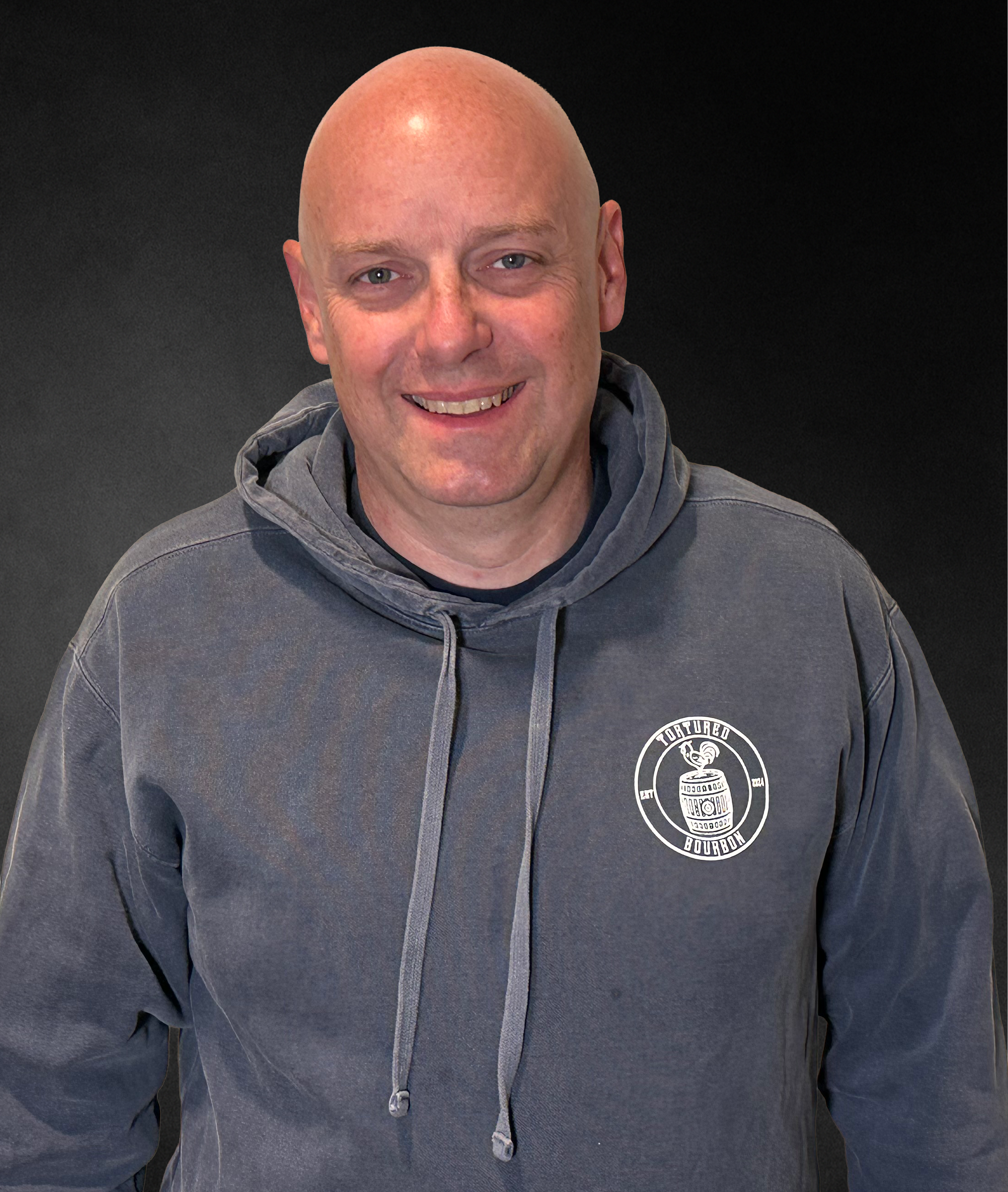A smiling man with a shaved head wearing a gray hoodie with a 'Tortured Bourbon' logo, standing against a dark background.