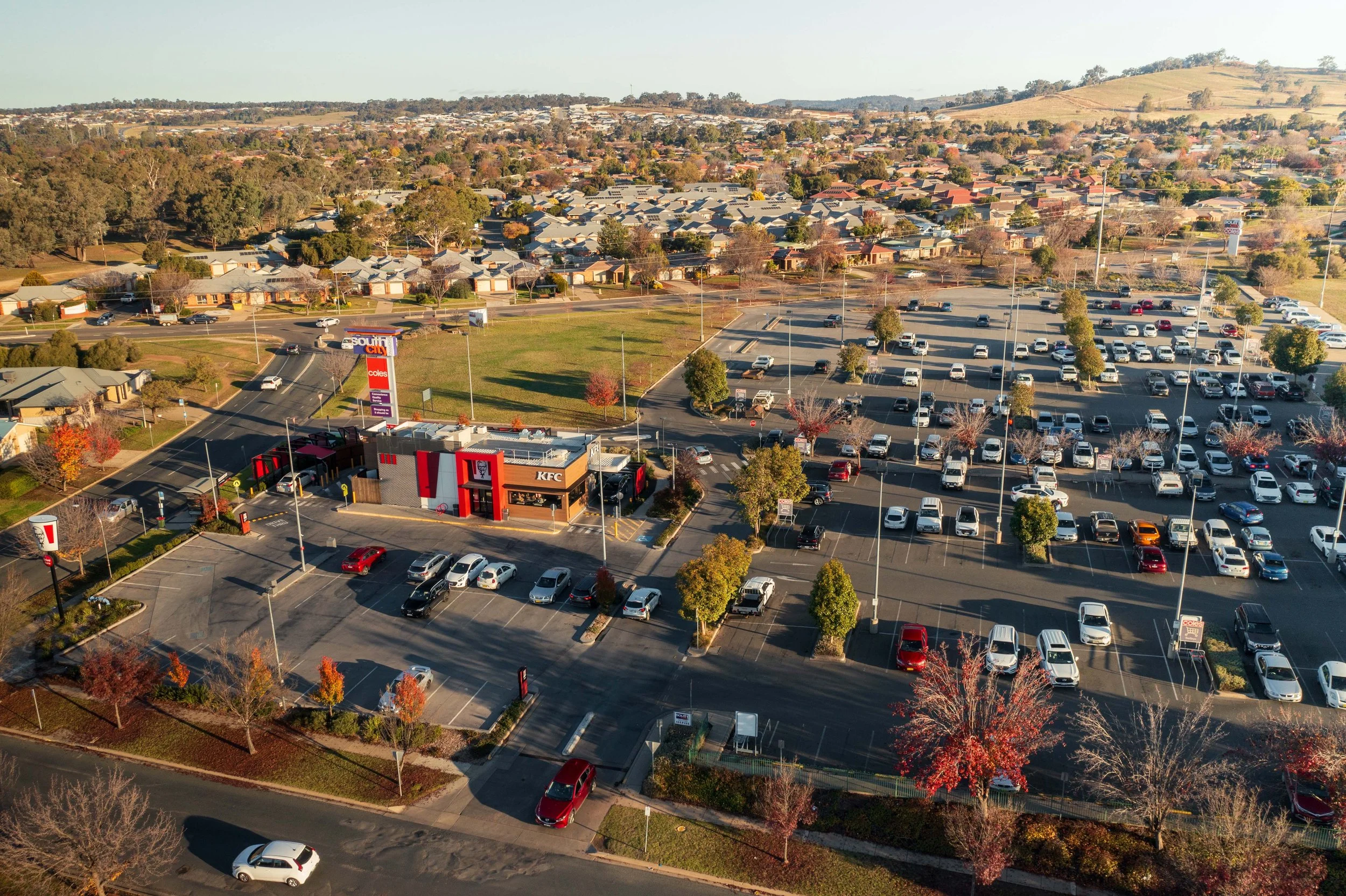 Aerial view of a shopping center parking lot with a KFC restaurant and South City sign, surrounded by residential neighborhoods and rolling hills in the background.
