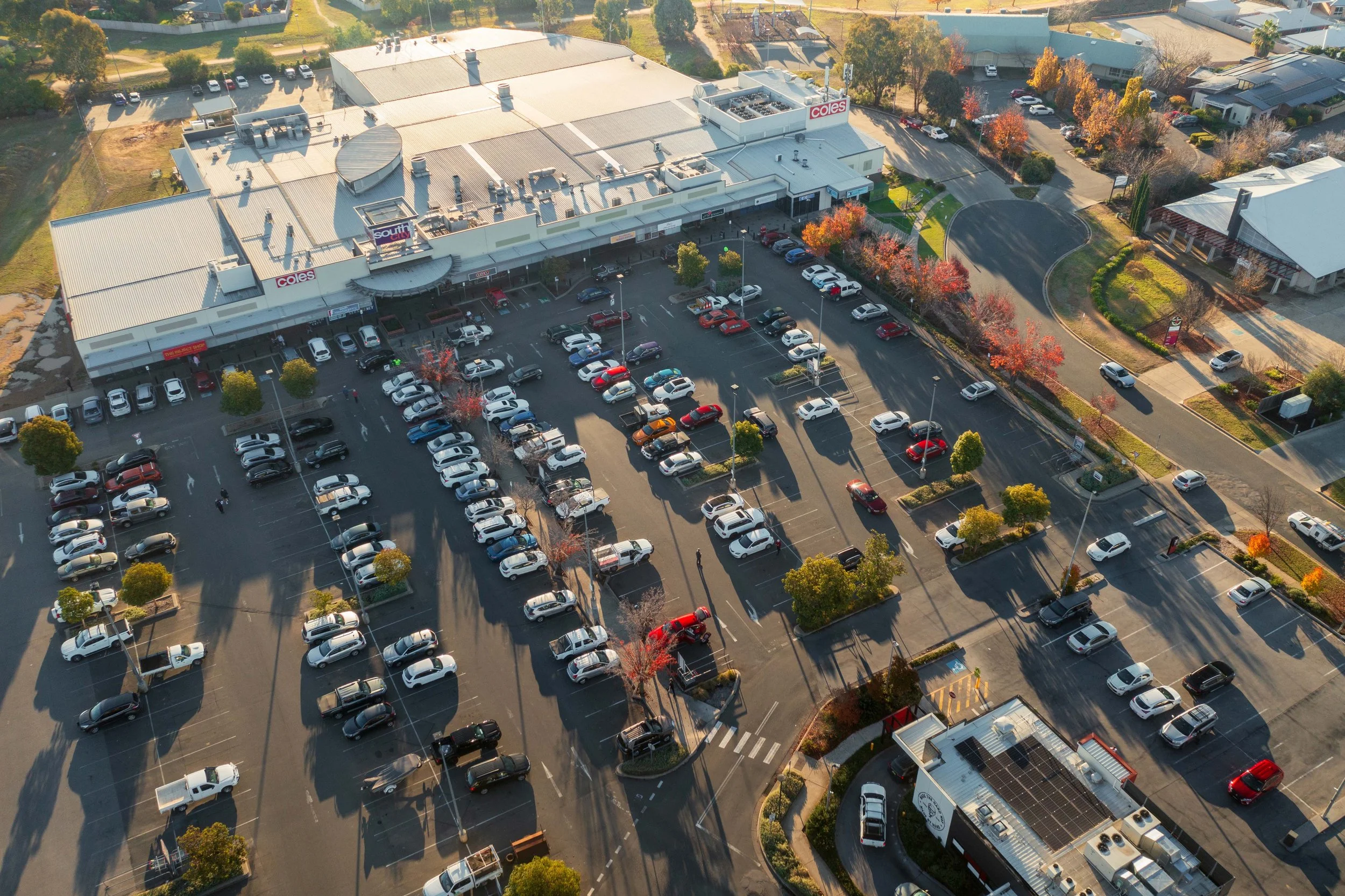 Aerial view of a retail shopping centre car park with several cars parked in marked spaces, surrounded by trees with autumn foliage, and a large retail store with a white roof and red signage at the front.