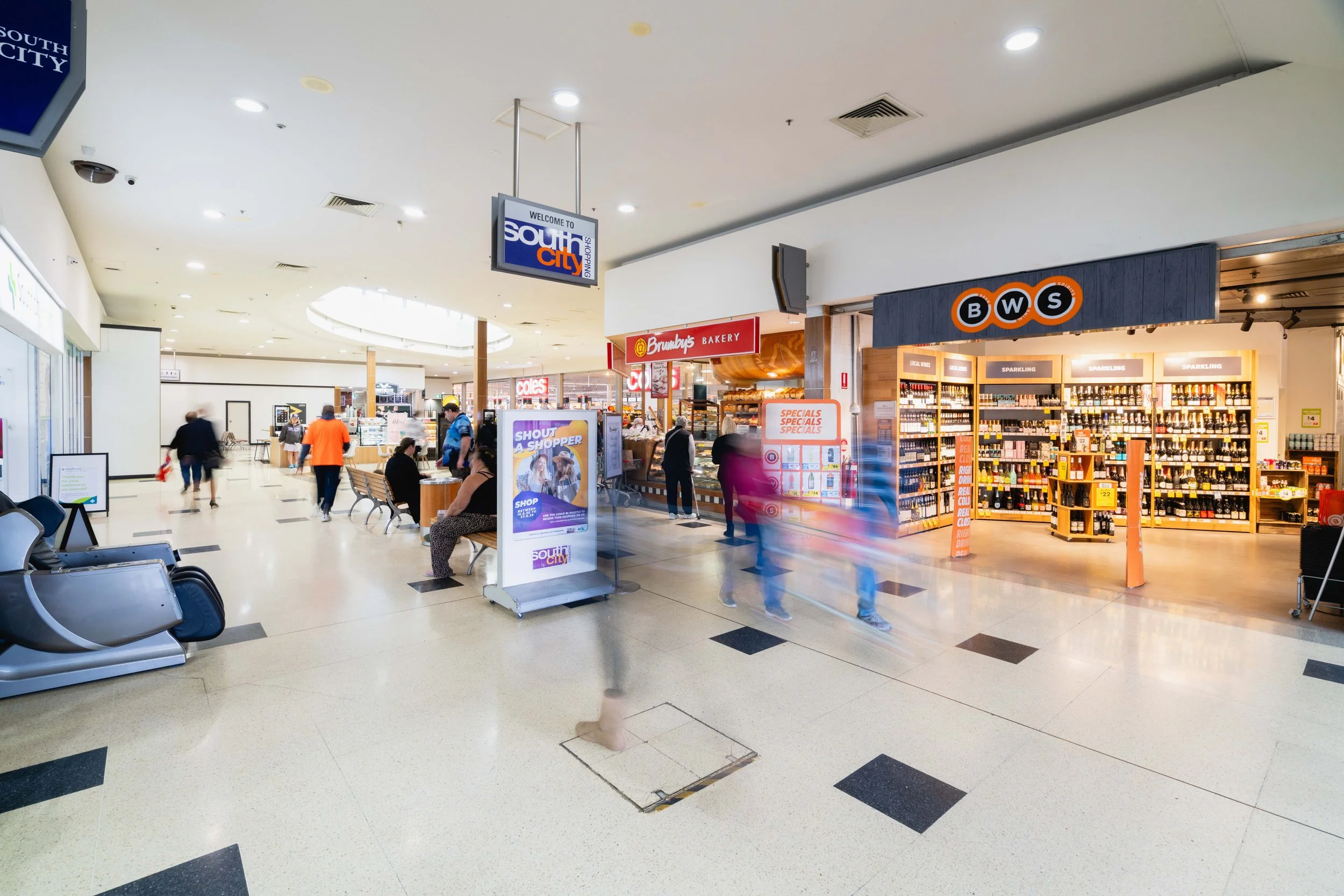 Interior of a shopping mall with people walking and sitting on benches; stores visible include a pharmacy and a wine shop, with a sign displaying "South City" overhead.