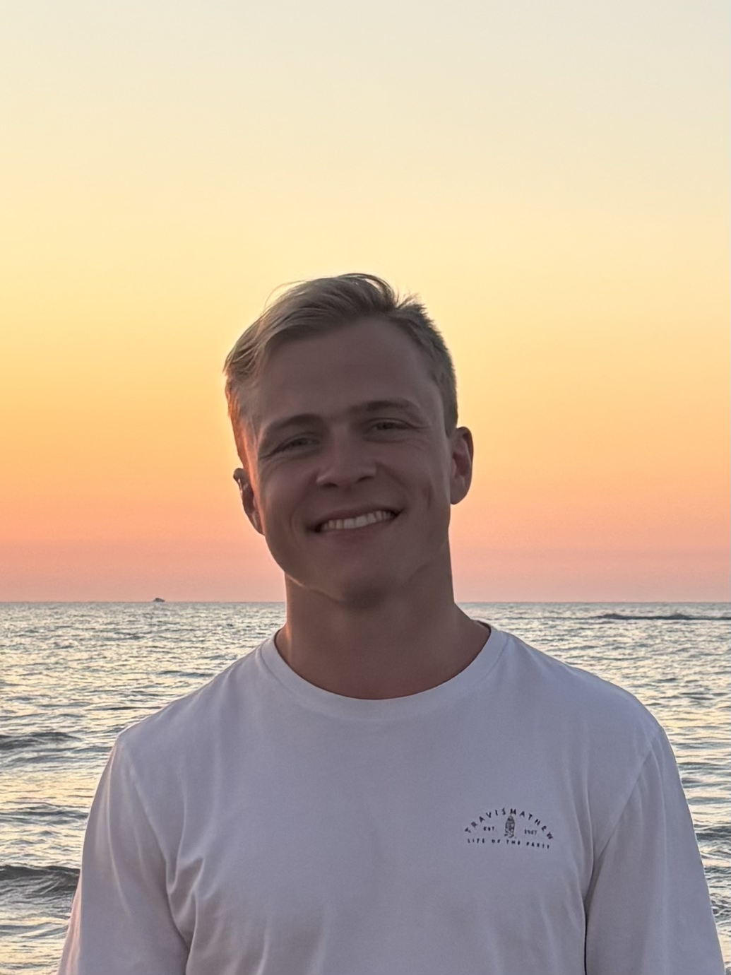 A young man smiling at the camera on the beach during sunset, with the ocean and a colorful sky in the background.
