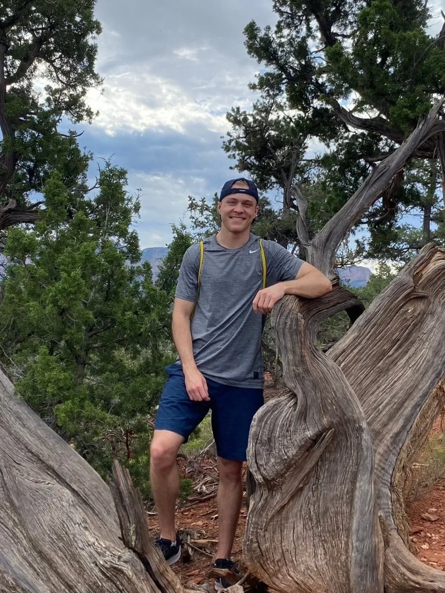 A young man standing outdoors, leaning on a twisted, weathered tree branch, surrounded by green trees and a cloudy sky.