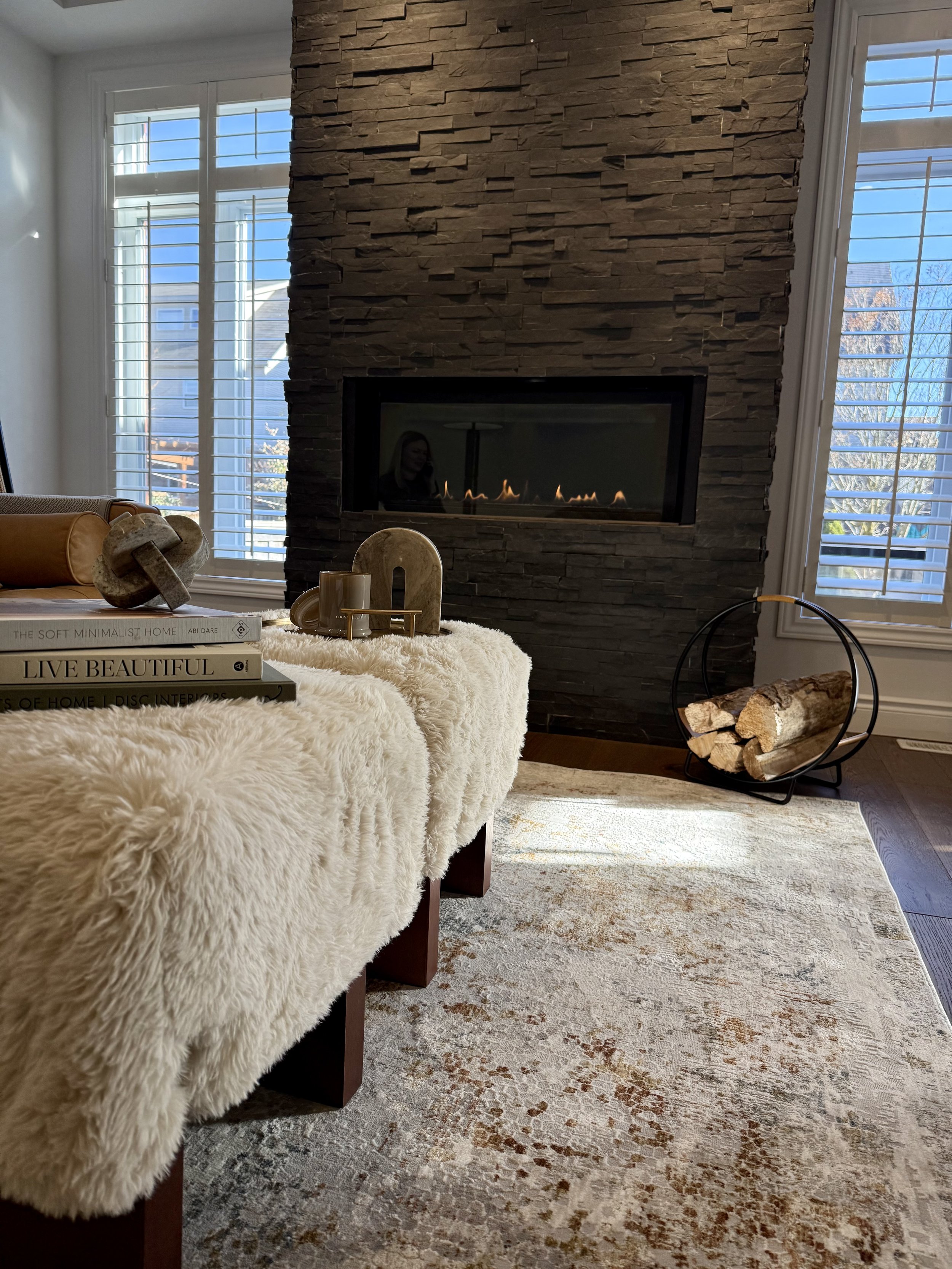 Black round dining table set with white plates, silver cutlery, and wine glasses, decorated with a clear glass vase with green foliage and white flowers, surrounded by four beige upholstered chairs, against a black and white floral wallpaper and a modern crystal chandelier hanging above.