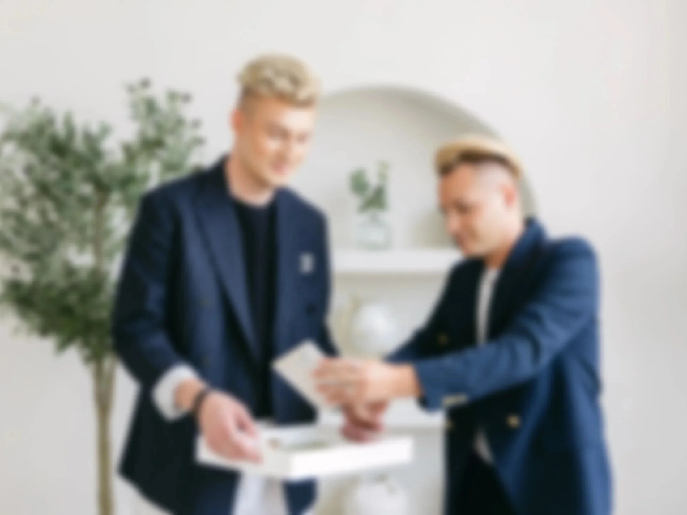 Two young men in formal suits exchange business cards in a bright, modern room with a round mirror and a potted plant in the background.