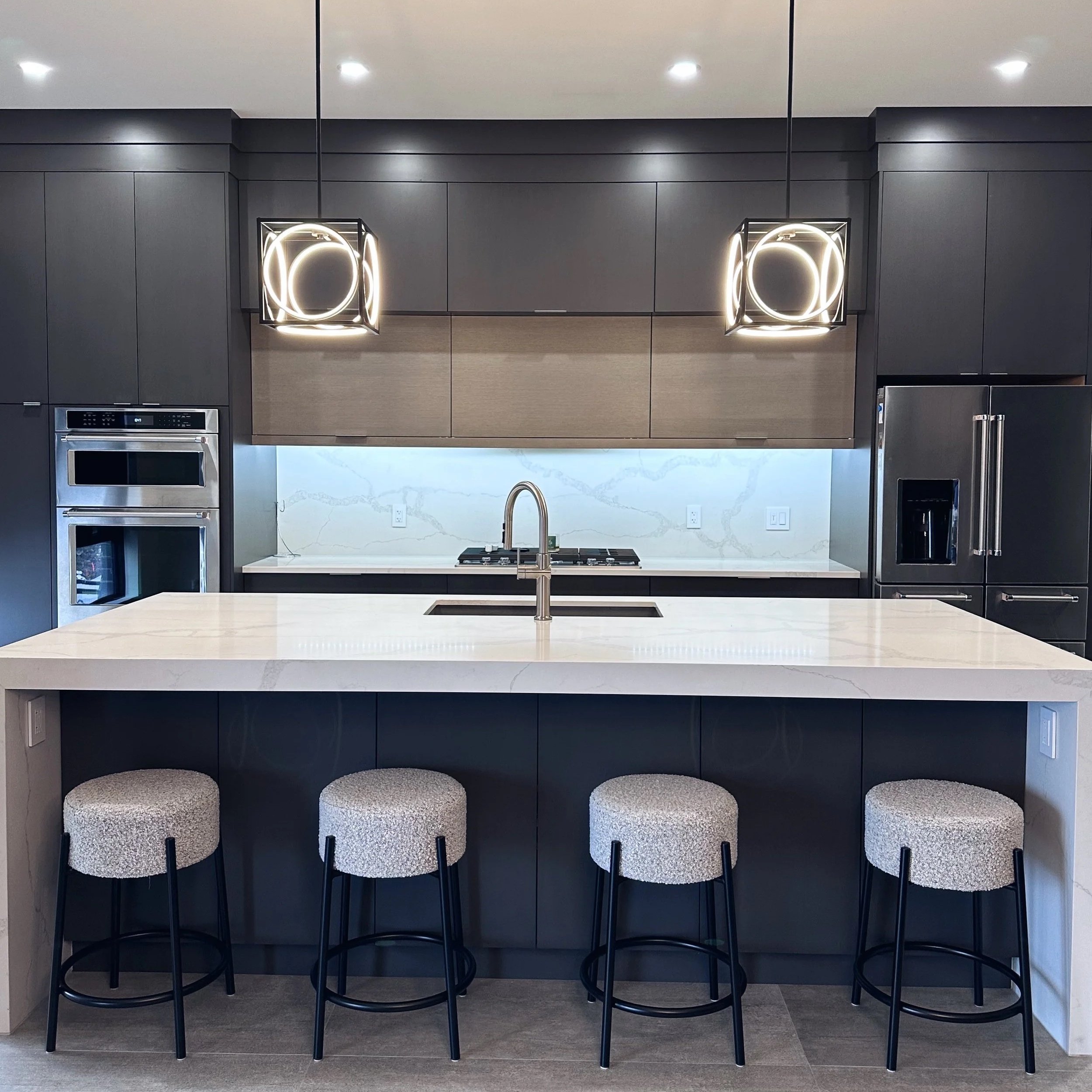 Modern kitchen with black cabinets, white marble island with seating, built-in stainless steel appliances, pendant lights, and a marble backsplash.