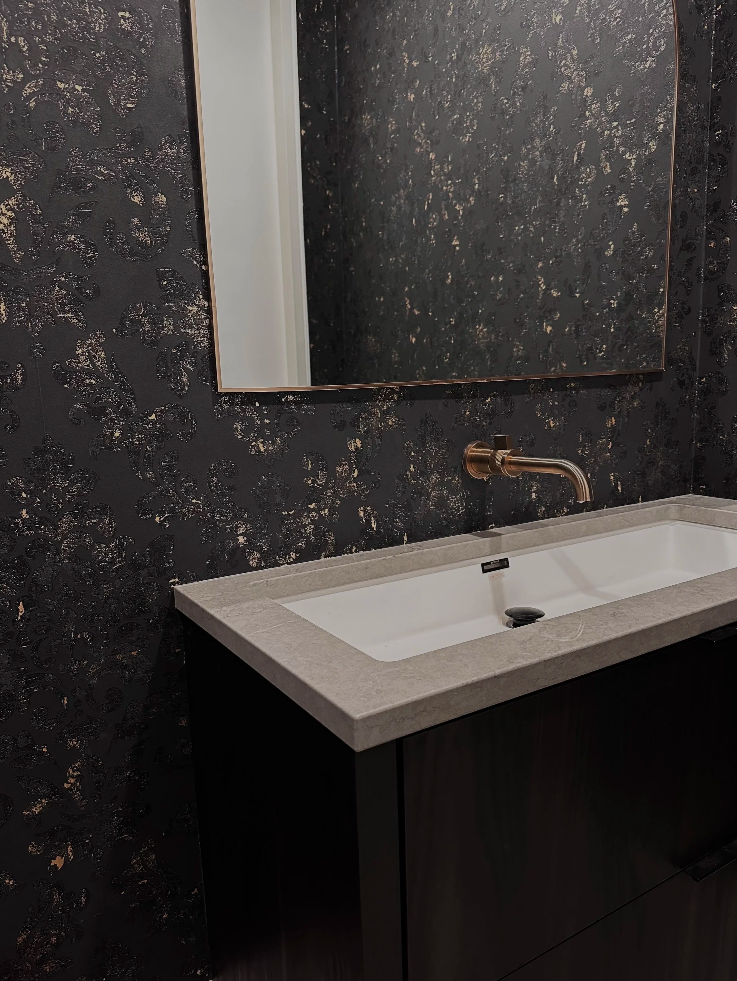 A modern bathroom sink with a brass faucet, a large mirror, and a black textured wallpaper with gold accents.