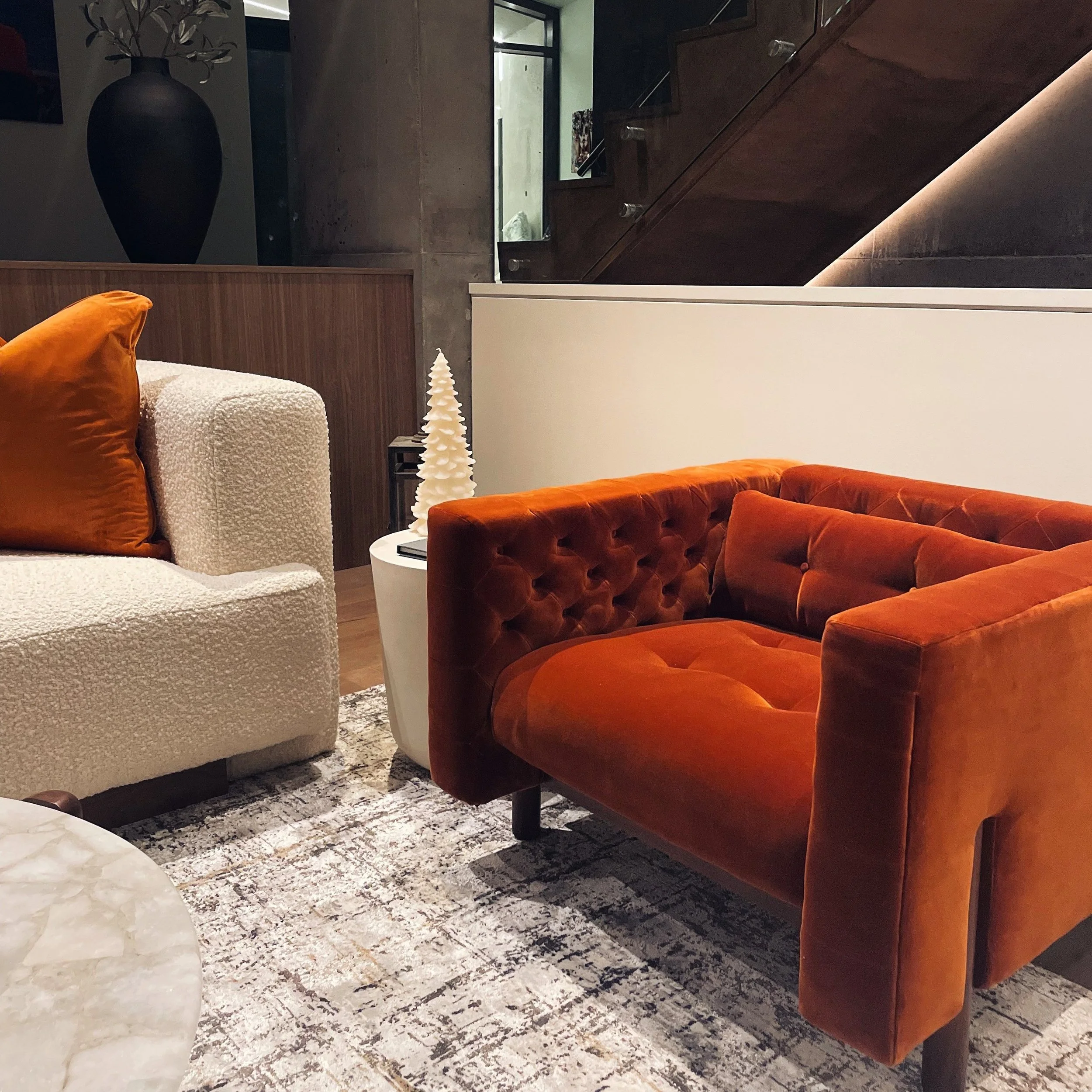 Living room with white textured sofa, orange velvet armchair, small white side table with a decorative Christmas tree, and a large dark vase in the background.