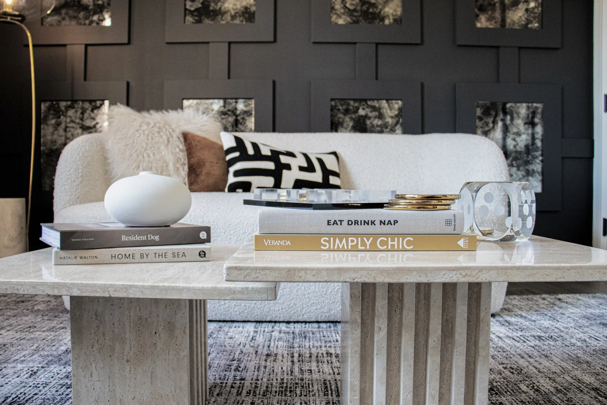 Living room coffee table with books, a decorative vase, and jewelry, in front of a sofa with pillows, against a dark wall with framed artwork.