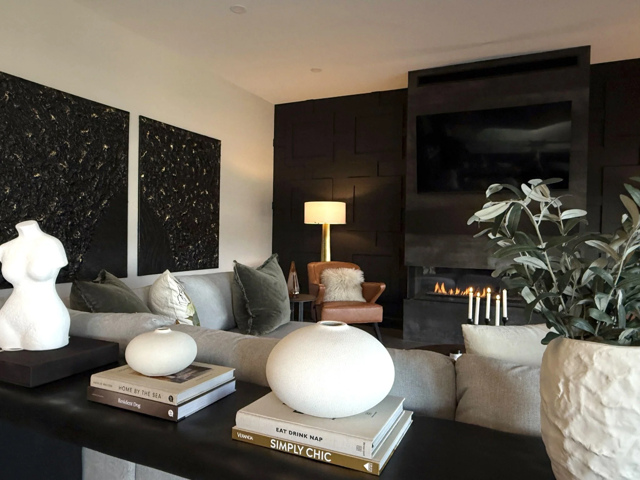 Modern living room with black textured wall art, beige sofa with pillows, a lamp, a leather armchair with a fur throw, a fireplace with candles, and a large potted plant.