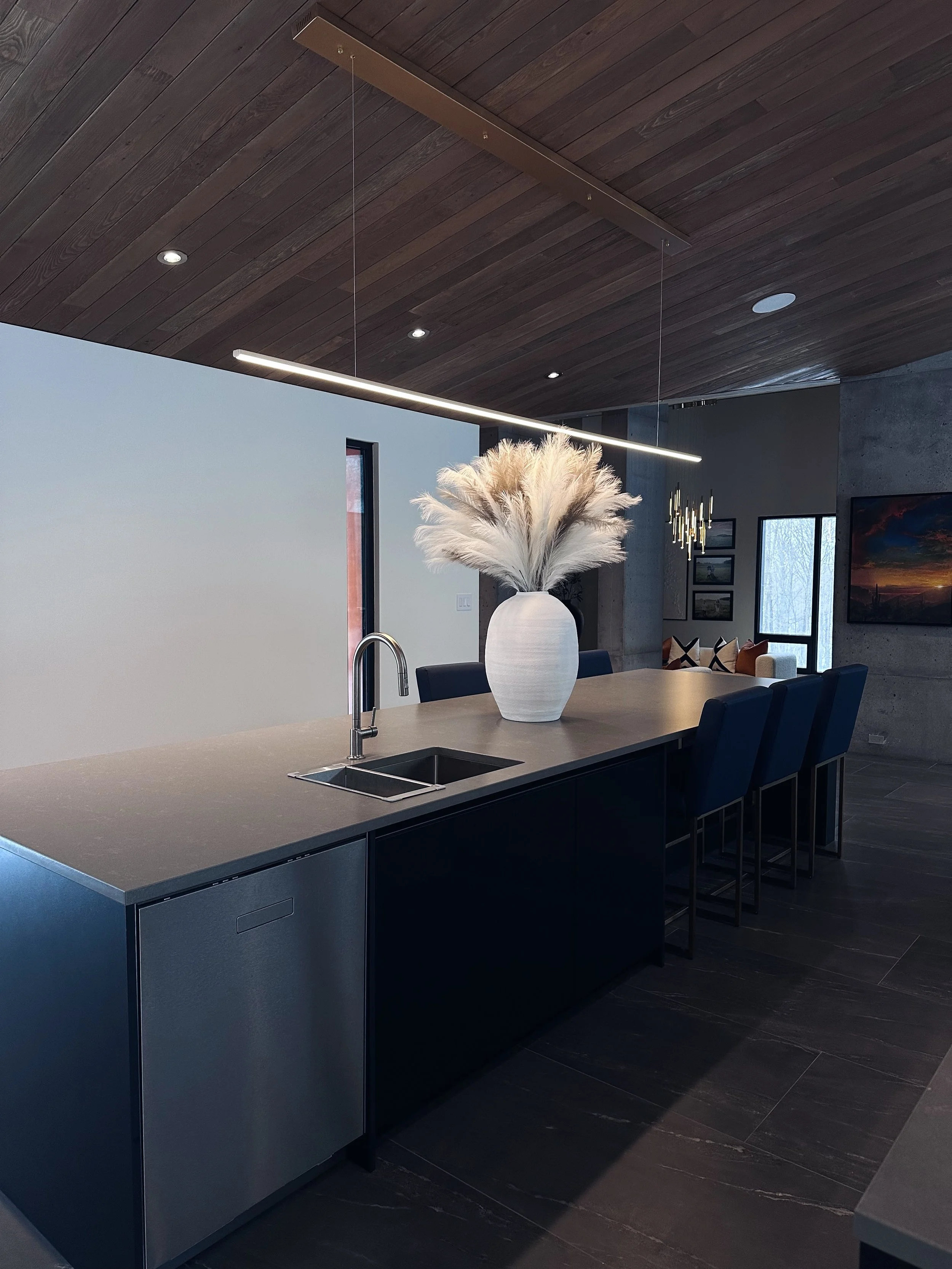 Modern kitchen island with a large white vase filled with pampas grass, dark cabinets, black chairs, a wooden ceiling with built-in lighting, and a concrete wall in the background.