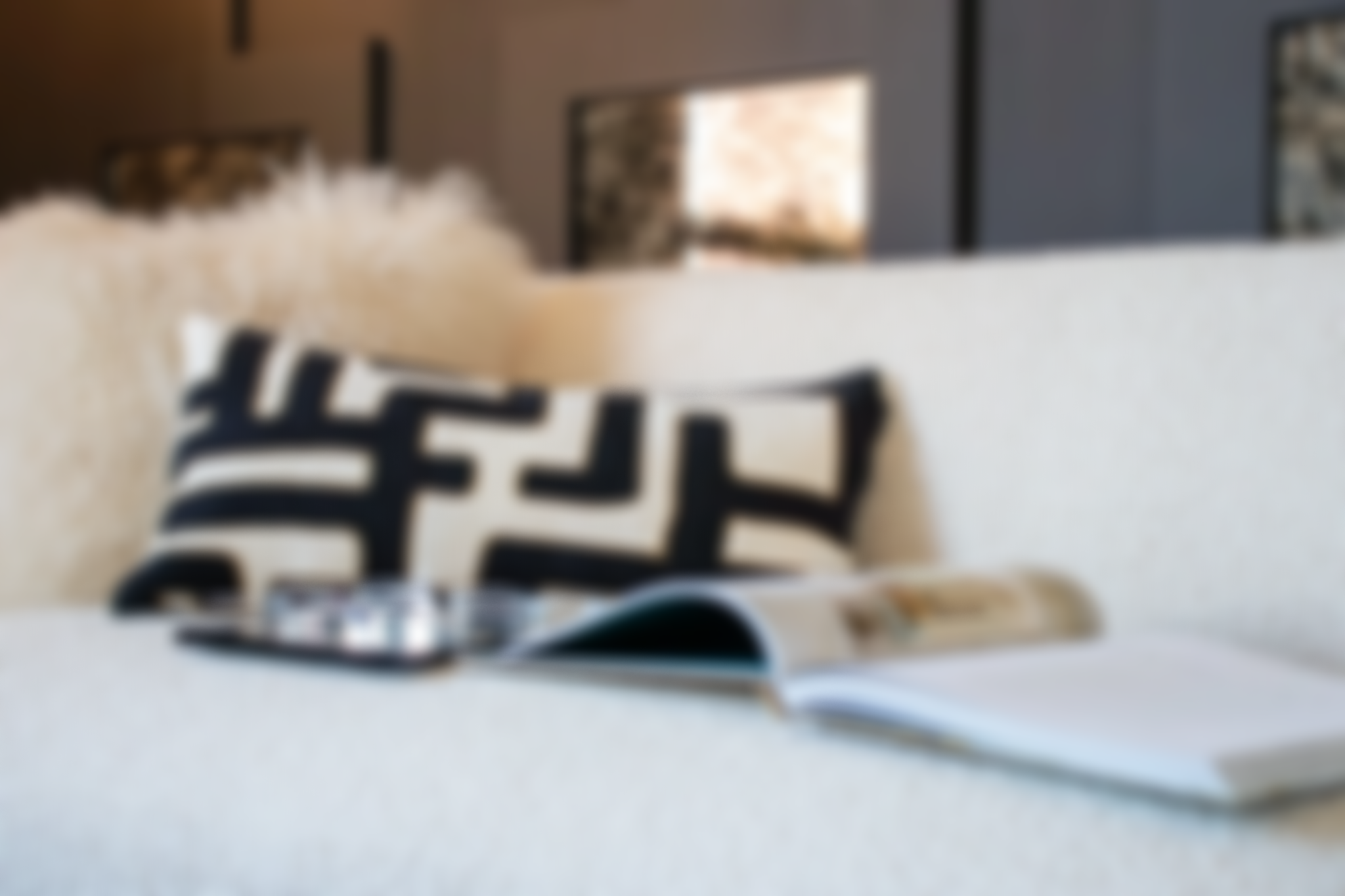 Blurred image of a cozy living room with a white sofa, black and white geometric patterned pillow, a tablet, and an open magazine on the sofa.